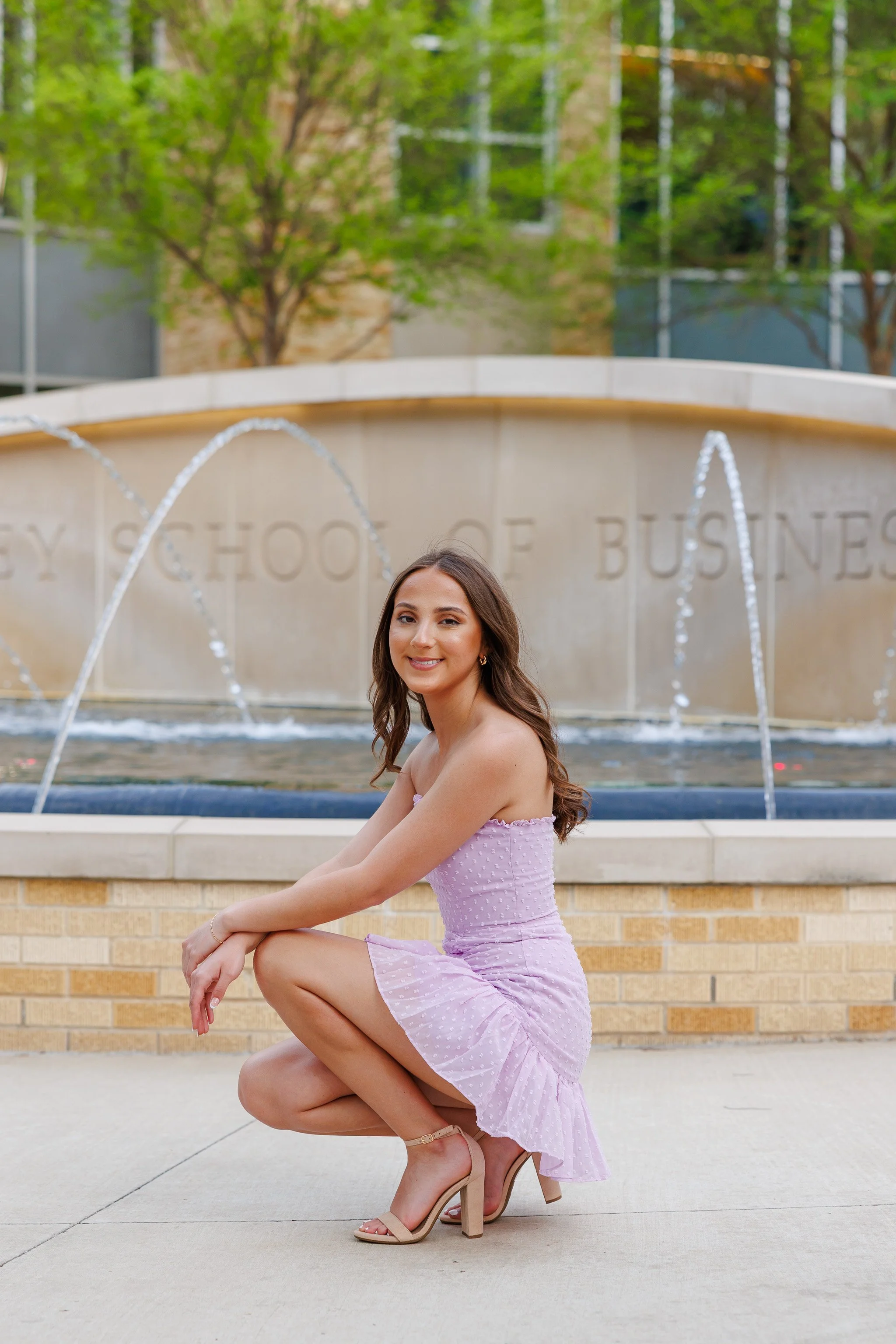 TCU Senior girl wearing lavender dress bending down in front of the water fountain at Neeley School of Business posing for senior pictures