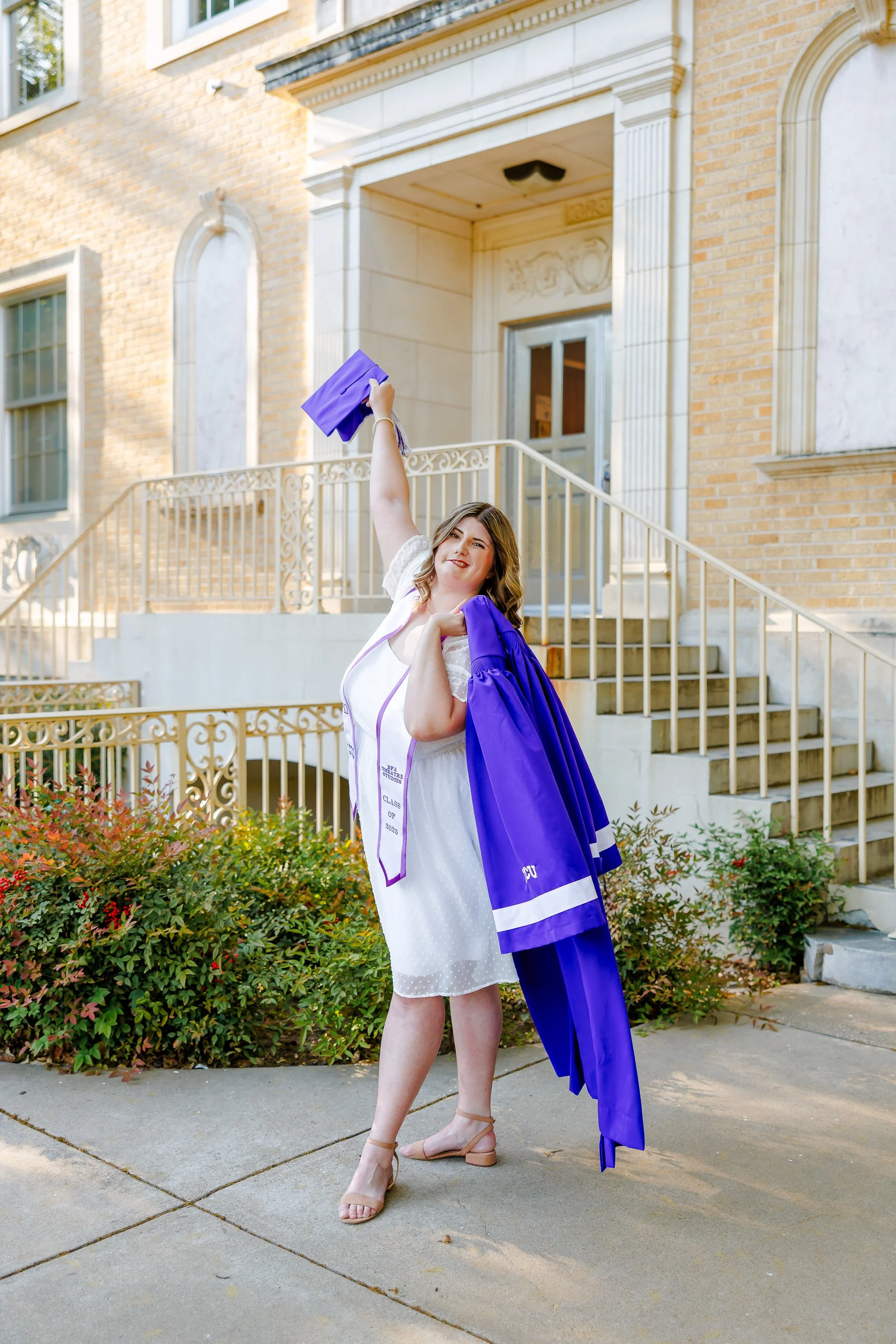 TCU senior girl in white dress holding a purple graduation gown over her shoulder and waving a graduation cap outside the Walsh Performing Arts Building for graduation portraits