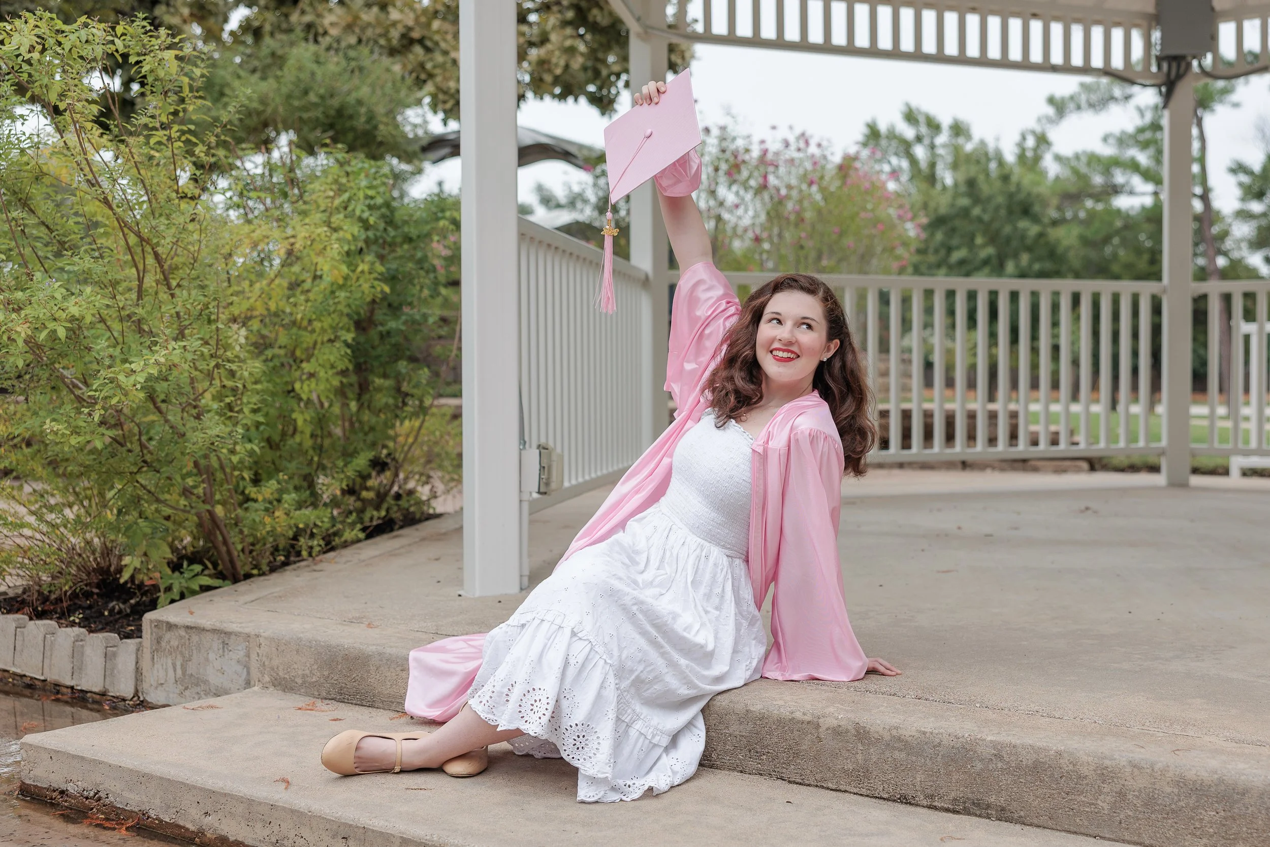 Woman in a pink graduation gown sitting on concrete steps beneath a gazebo, smiling while holding her matching cap in the air, showcasing graduation photos with a pose naturally filled with excitement and pride.