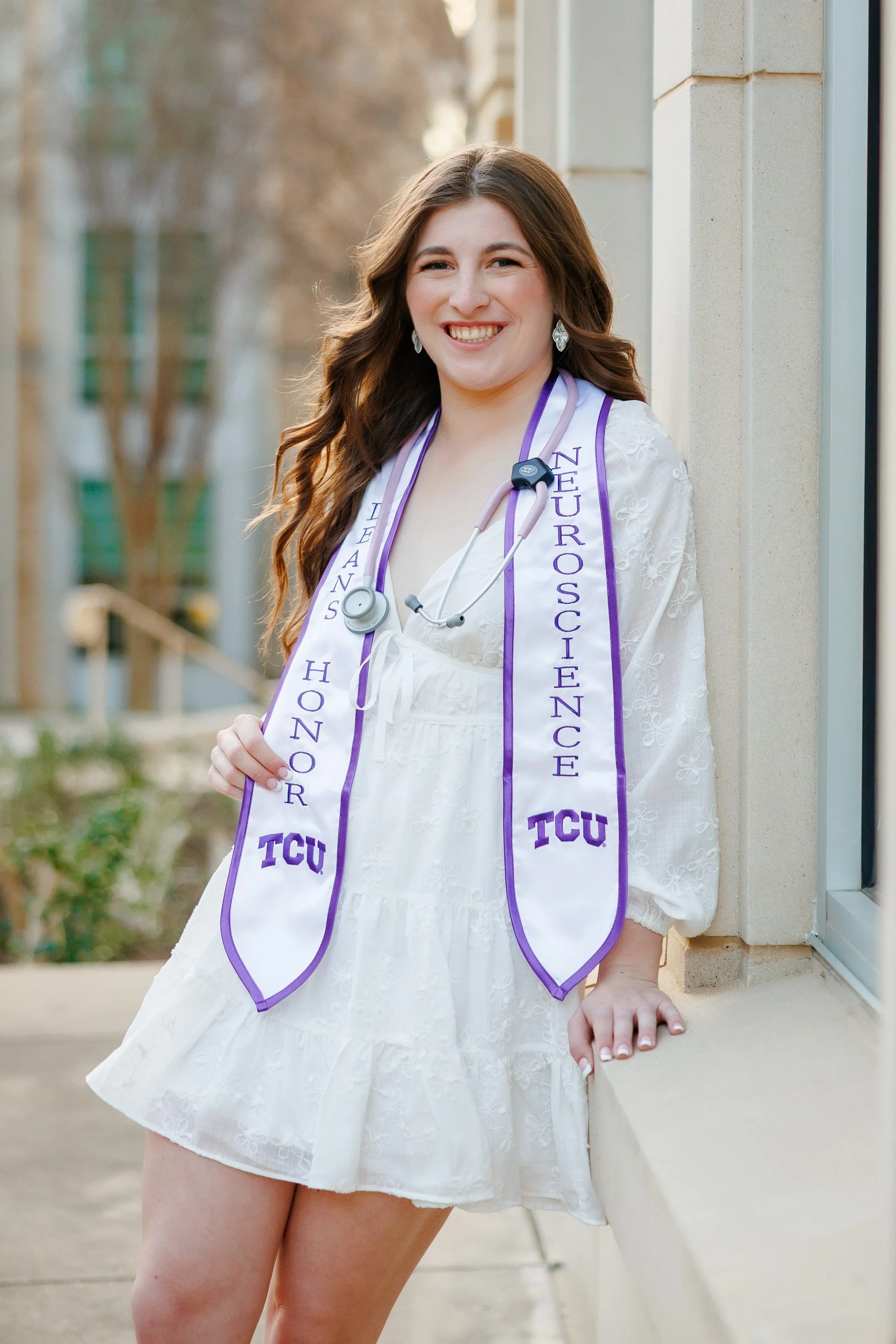 Senior girl wearing white dress walking with a neuroscience stole near Scharbauer Hall at TCU posing for senior pictures