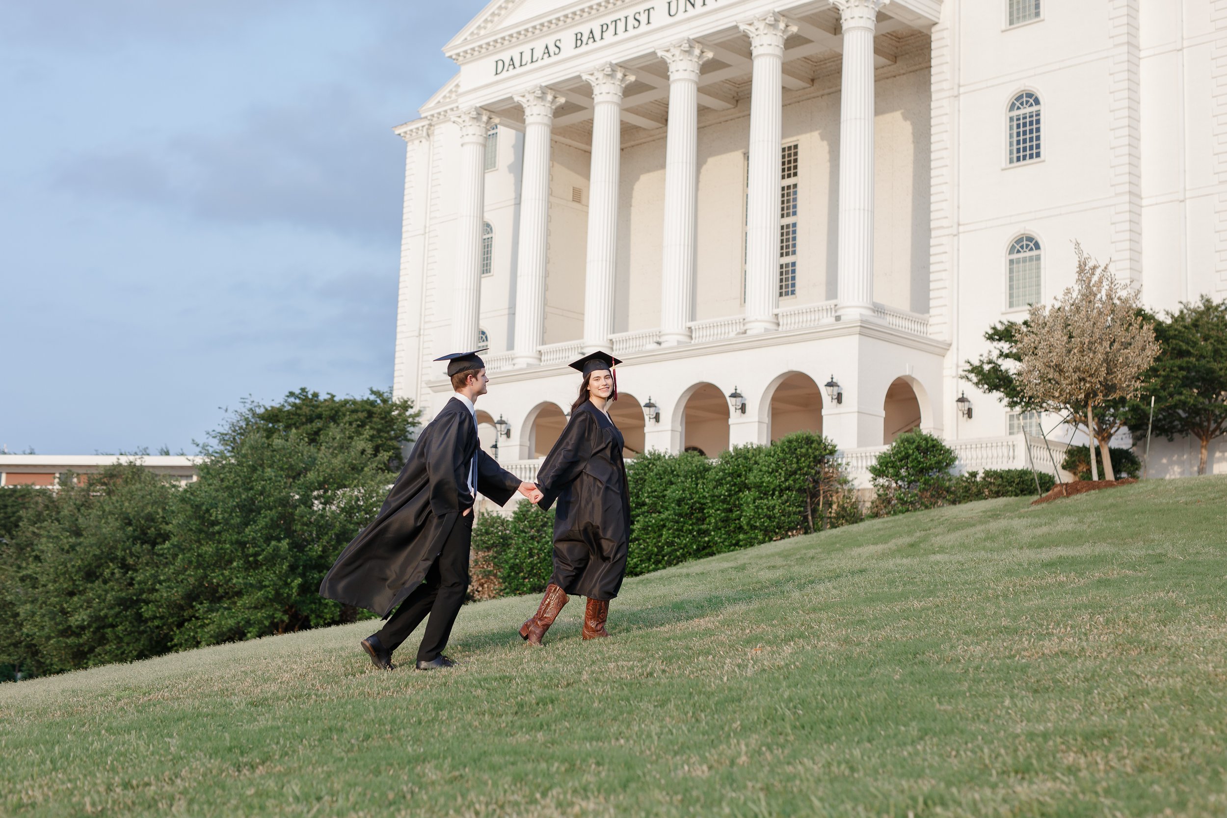 Graduating couple in caps and gowns holding hands while walking up a grassy hill in front of the Dallas Baptist University building with white columns, captured for couples graduation photos by a Dallas photographer.