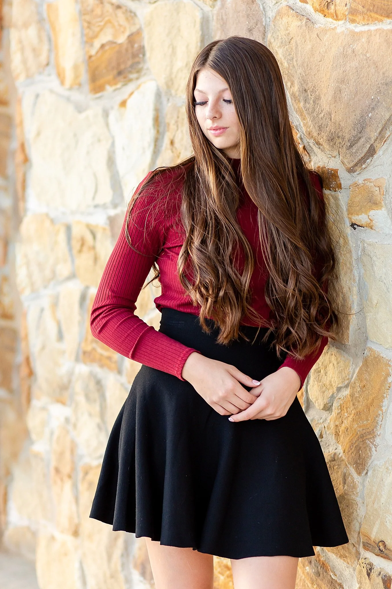 young lady with very long brown wavy hair wearing a maroon turtleneck short black skirt with eyes shifted toward the ground leaning against a rock wall posing for senior pictures in Colleyville