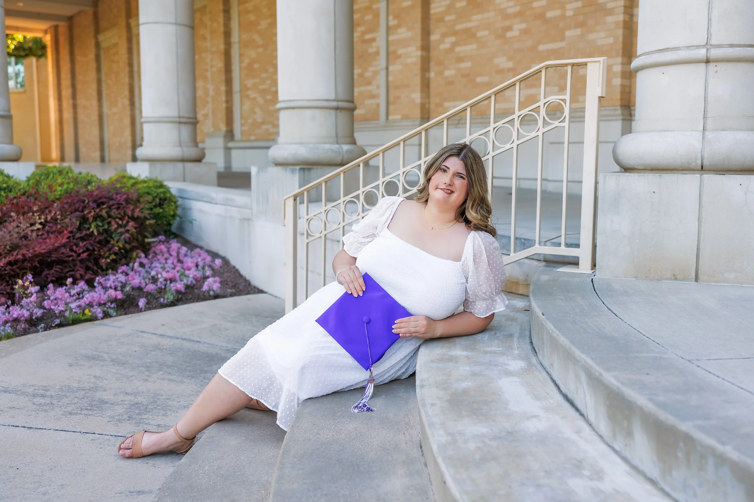 TCU senior girl in white dress sitting on steps holding a purple graduation cap outside the Walsh Performing Arts Building for graduation portraits