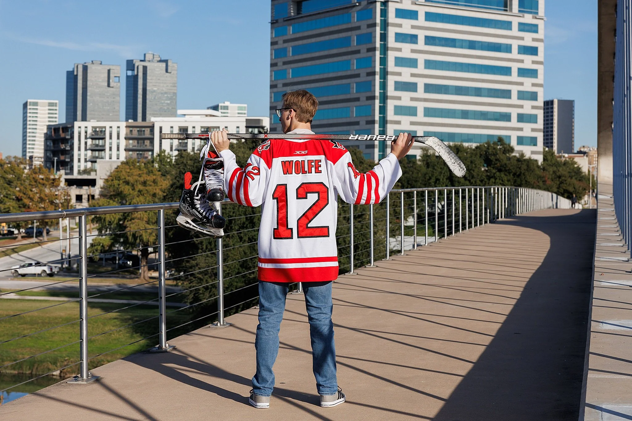 young man wearing jeans and a red #12 hockey jersey holding a hockey stick with skates hanging off looking away from the camera standing on 7th street bridge posing for senior pictures
