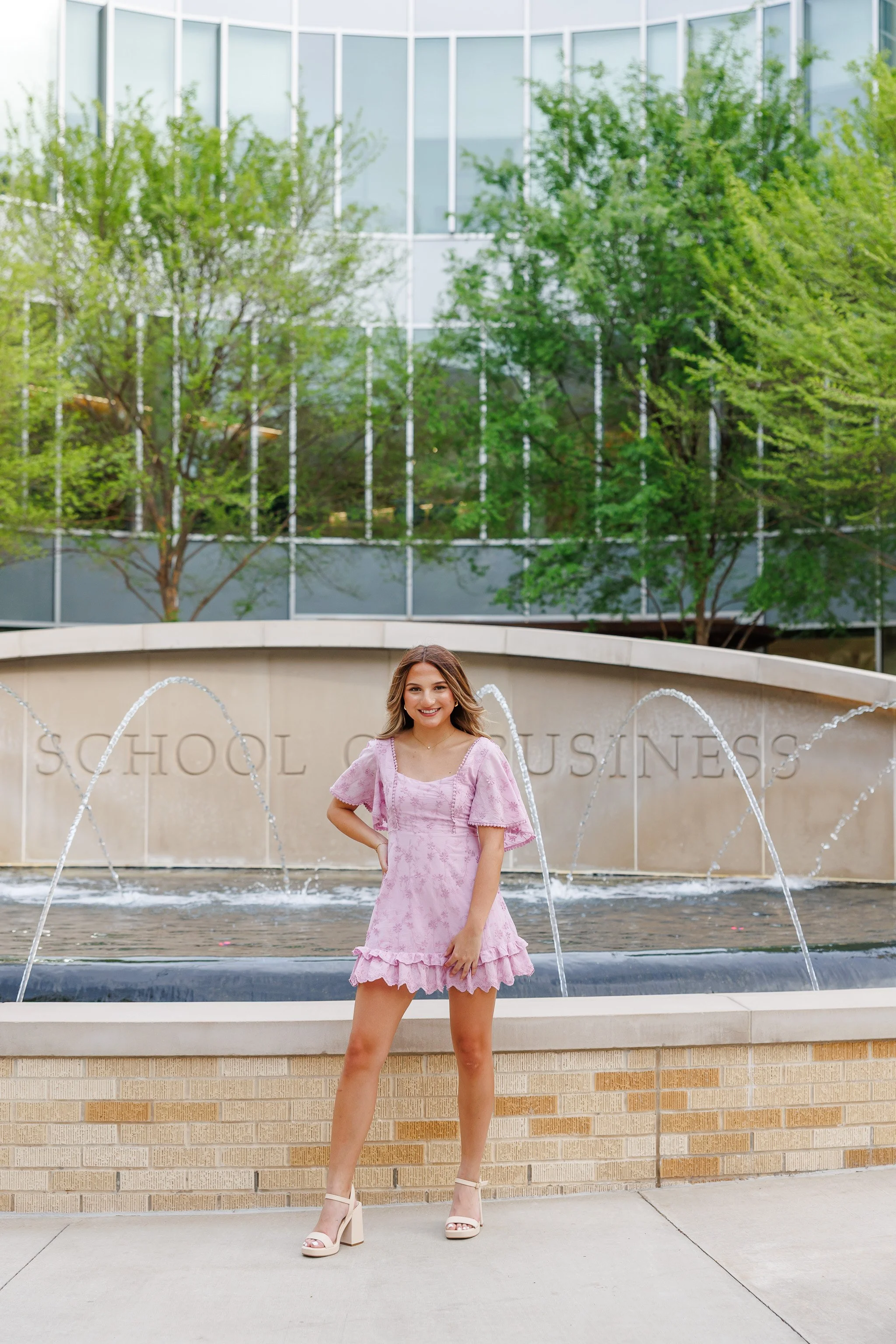 TCU Senior girl wearing lavender dress standing in front of the water fountain at Neeley School of Business posing for senior pictures
