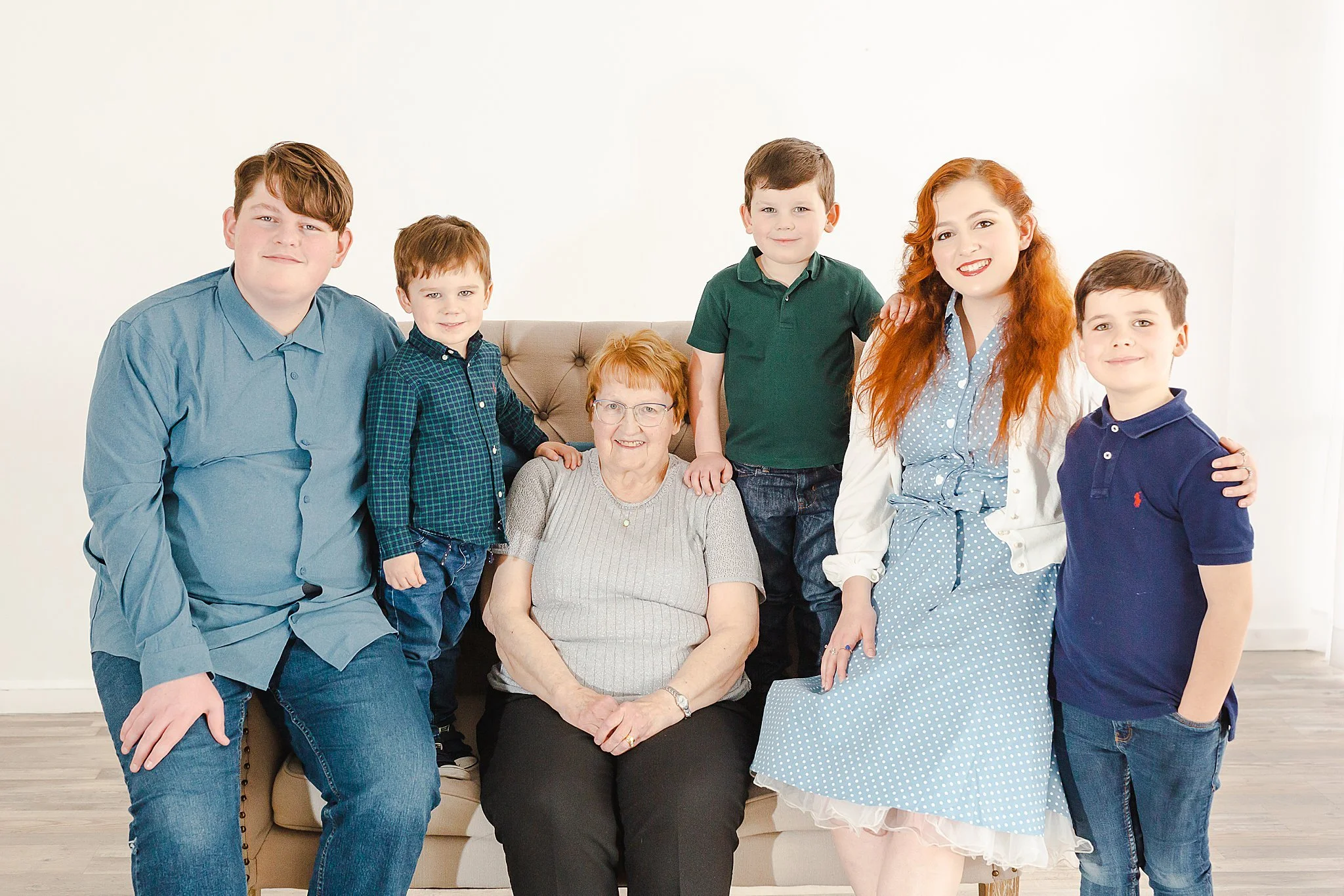 five grandkids in nice shirts and blue jeans surrounding their grandmother who is sitting on a coach smiling at the camera posing for professional portraits at New Light Studio Bedford Texas