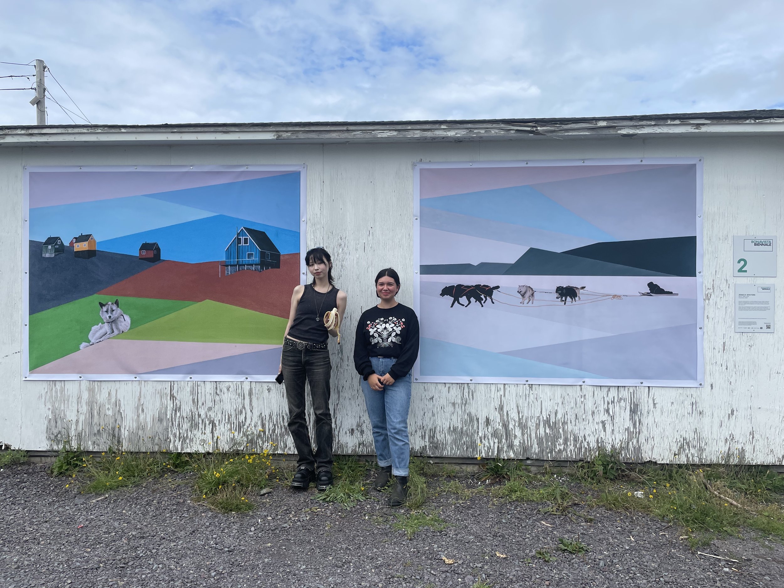  Megan Kyak-Monteith and Ooleepeeka Eegeesiak stand in front of Jessica Winter’s mural,  Hopedale , during the Bonavista Biennale, August 2023. Bonavista, NL. Photograph by Danielle Miles. 