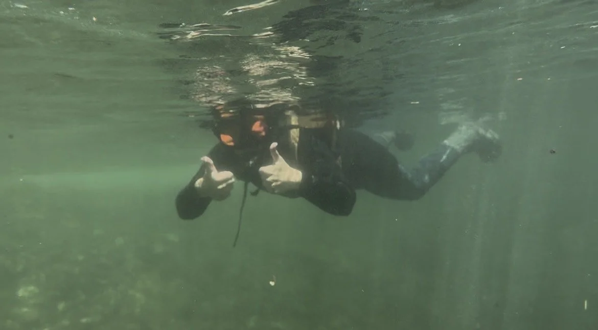A snorkeler offers two thumbs-up to the camera while swimming underwater. She wears a wetsuit, backpack, and snorkel mask.