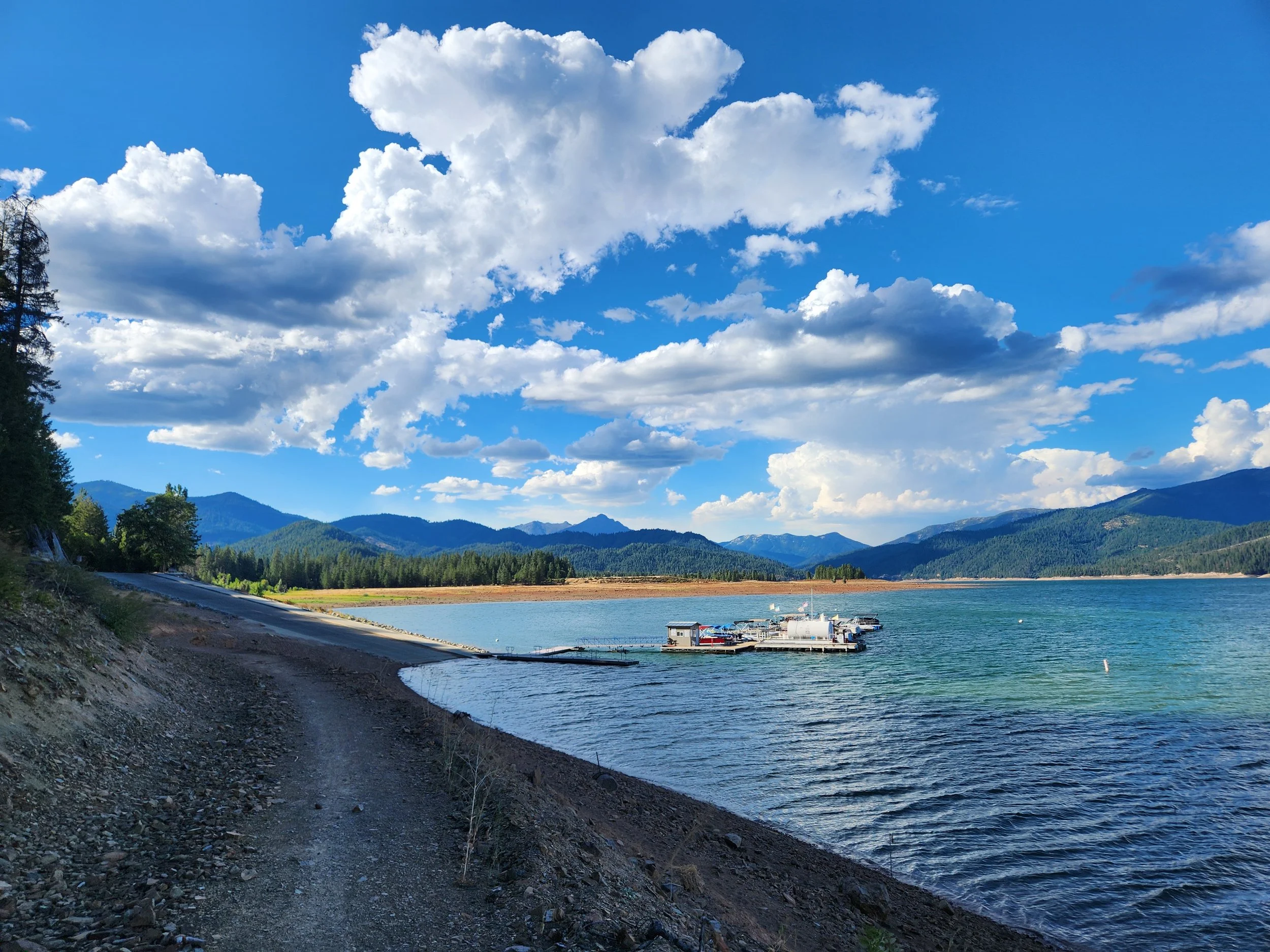 Lake with a small dock and boat, surrounded by mountains and forest, under a blue sky with large white clouds.
