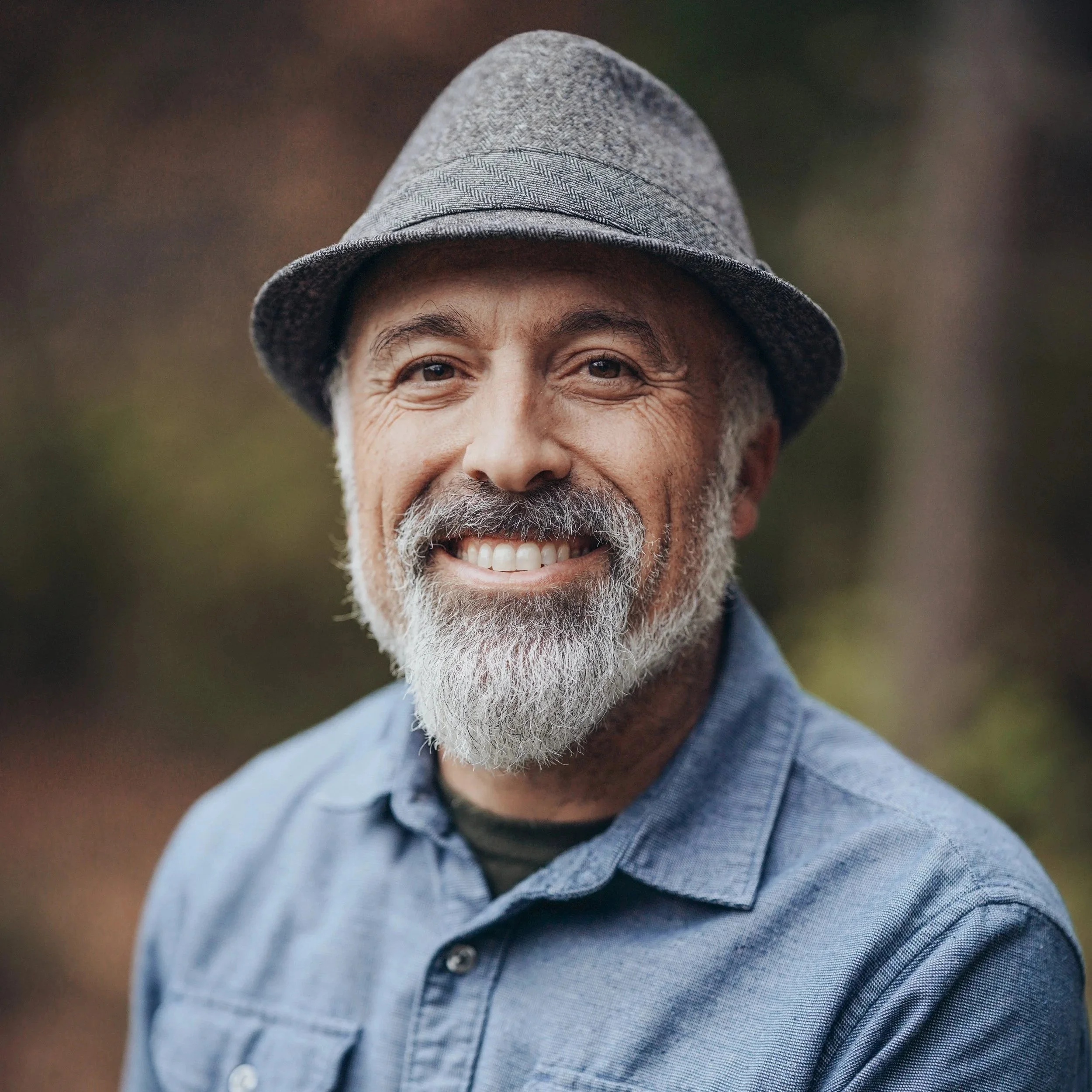 Nick Goulette smiles at the camera. He wears a felt hat and blue button down shirt. He has a salt and pepper beard and mustache.