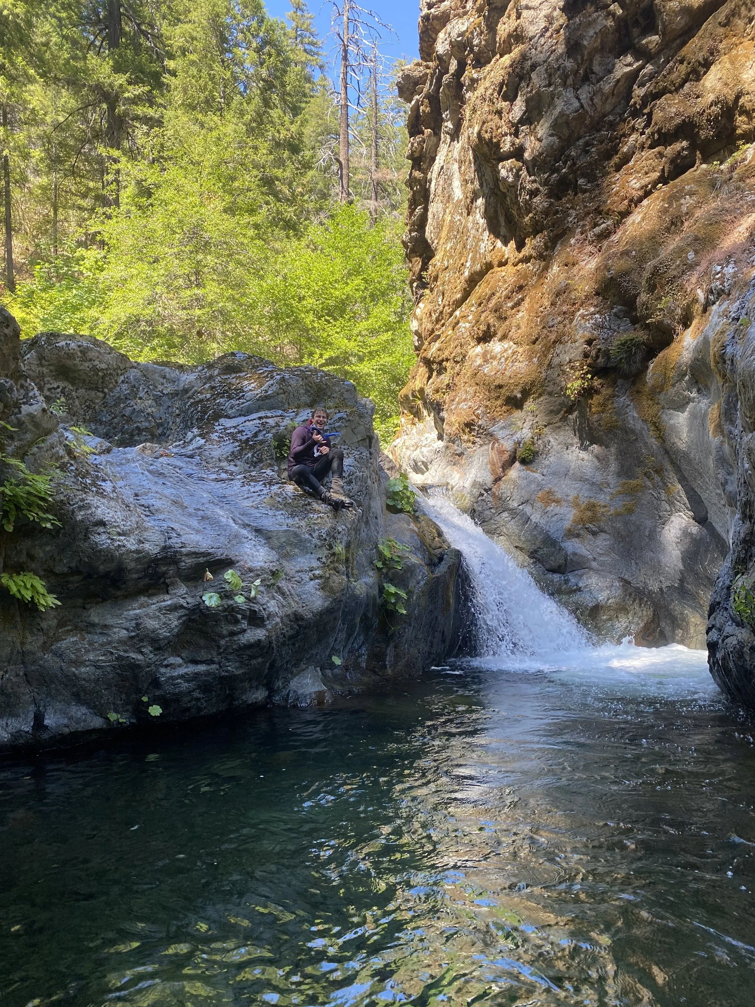 A fisheries technician sits on a large boulder next to a small waterfall. He has removed his snorkel mask to smile at the camera.