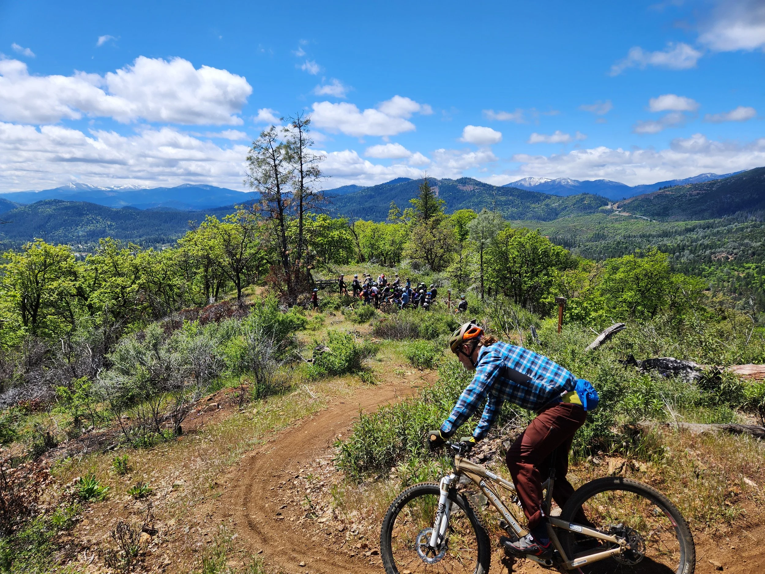 A person mountain biking on a dirt trail through a lush green forest with a large group of cyclists ahead. The background features distant mountains under a partly cloudy sky.