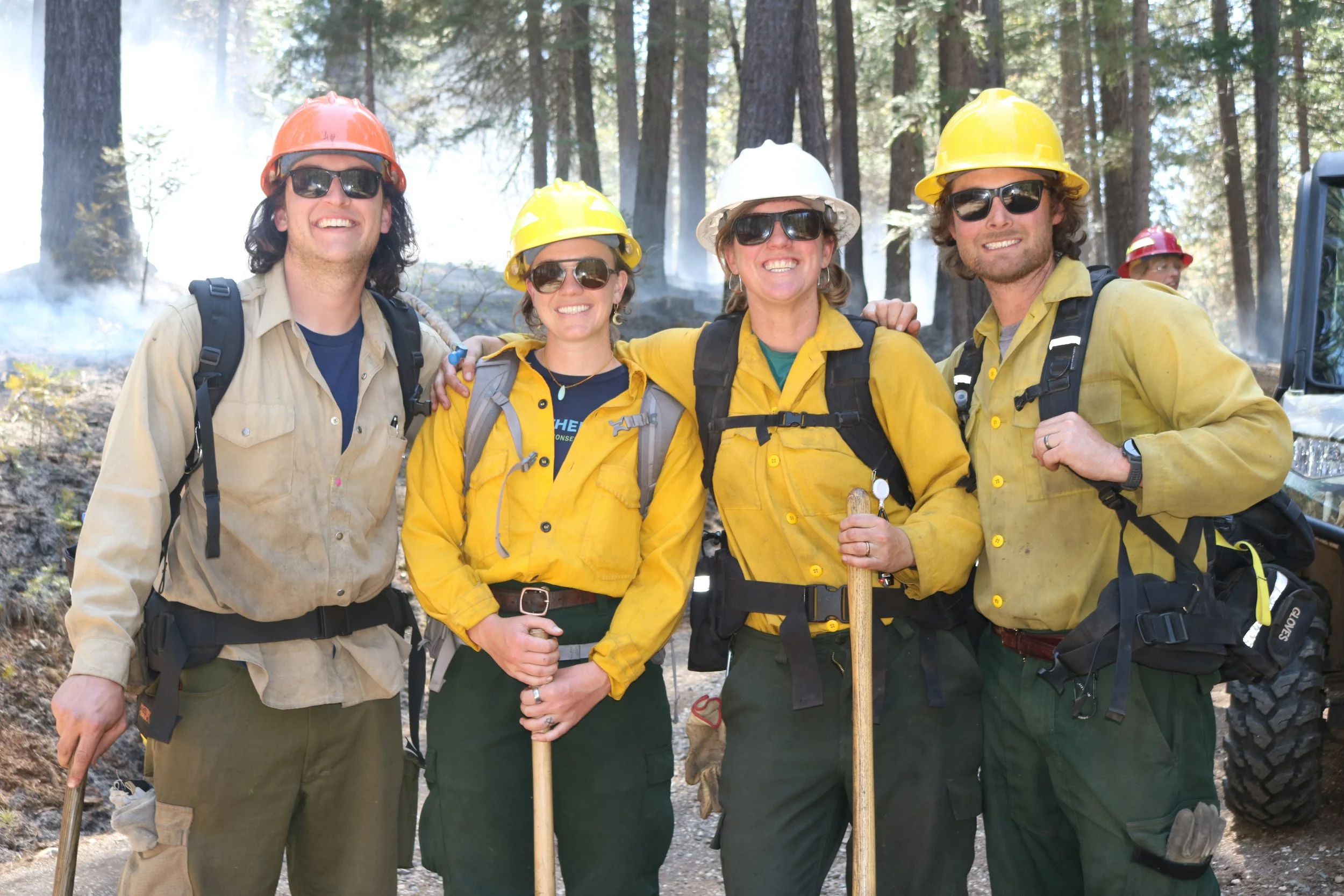 Two men and two women dressed in fire-appropriate clothing and gear smile at the camera. They wear hard hats, sunglasses, fire packs, and hold hand tools.