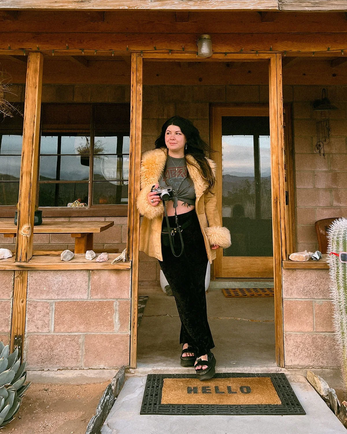 Woman designer with long brown hair standing on desert cabin porch holding camera, wearing fur-trim coat, band tee, and platform sandals