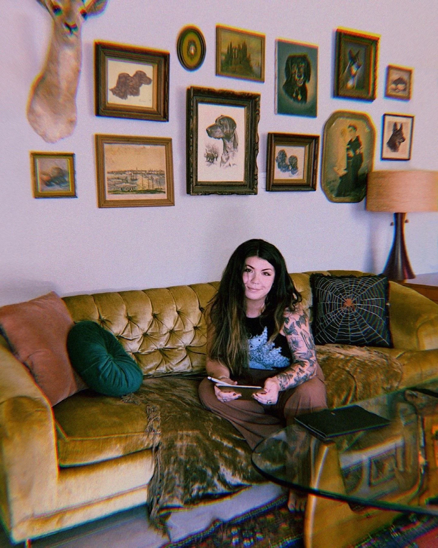 Tattooed woman designer with long brown hair sitting on a gold velvet couch with gallery wall of eclectic vintage artwork