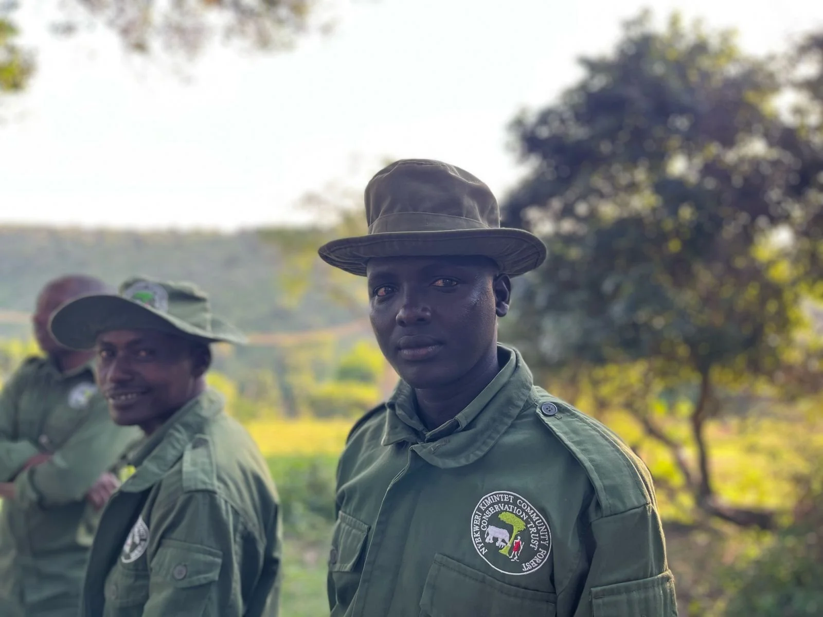 Three men in green uniforms with patches, outdoors in a green, wooded area. The man in the foreground has a serious expression and is wearing a wide-brimmed hat. The other two men are in the background, with one smiling and wearing a similar hat.