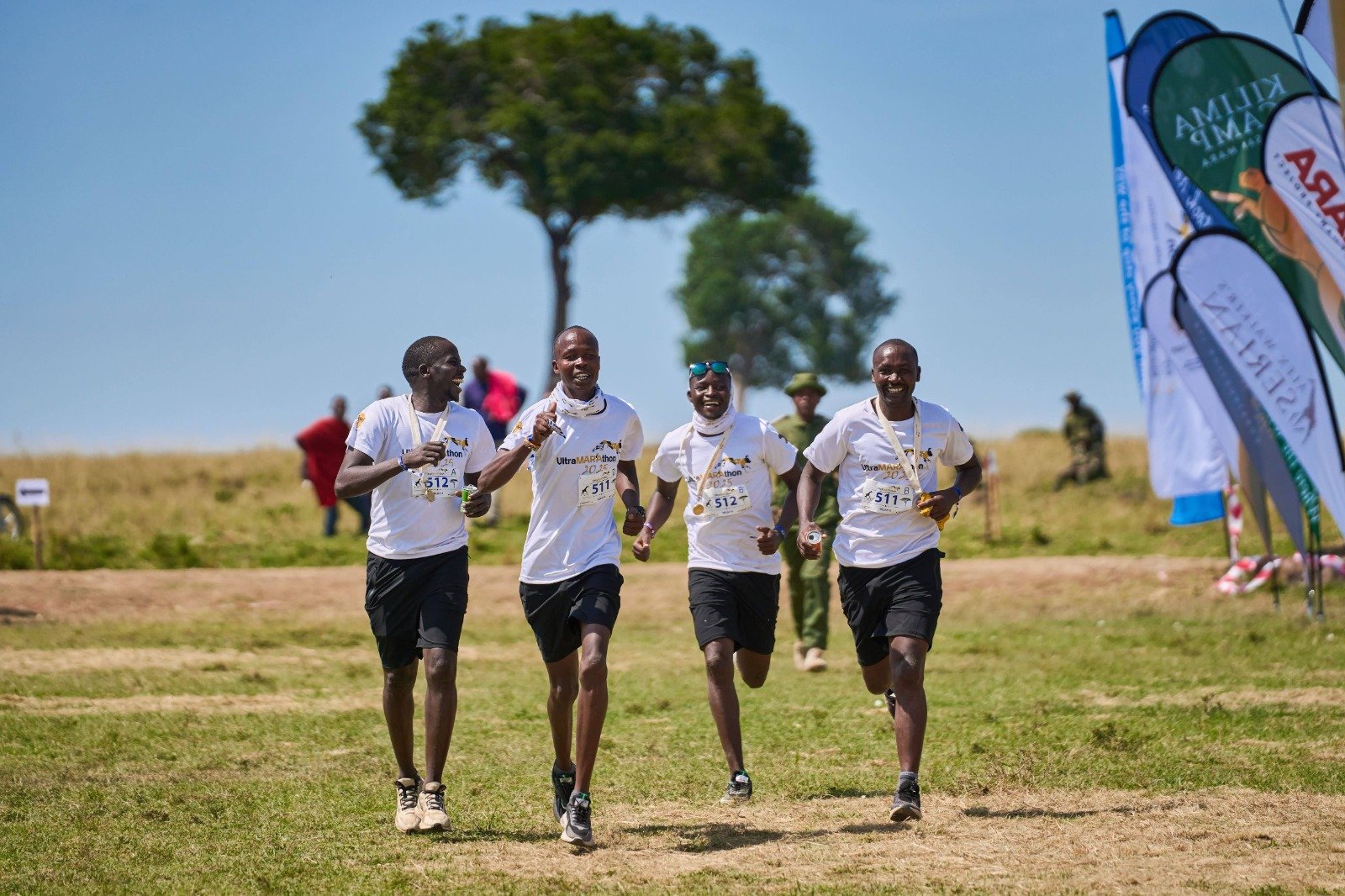A woman with braided hair smiling outdoors in a park or garden, wearing a gray vest over a blue shirt with rolled-up sleeves, a watch on her left wrist, and a multicolored beaded bracelet on her right wrist.