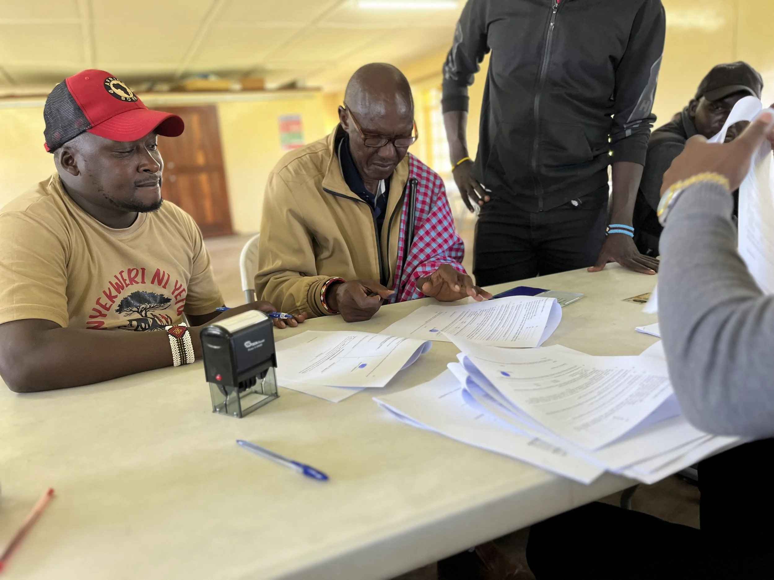 A group of men sit around a table looking at documents, with some standing. They are engaged in a discussion or review, with papers and a stamp on the table.