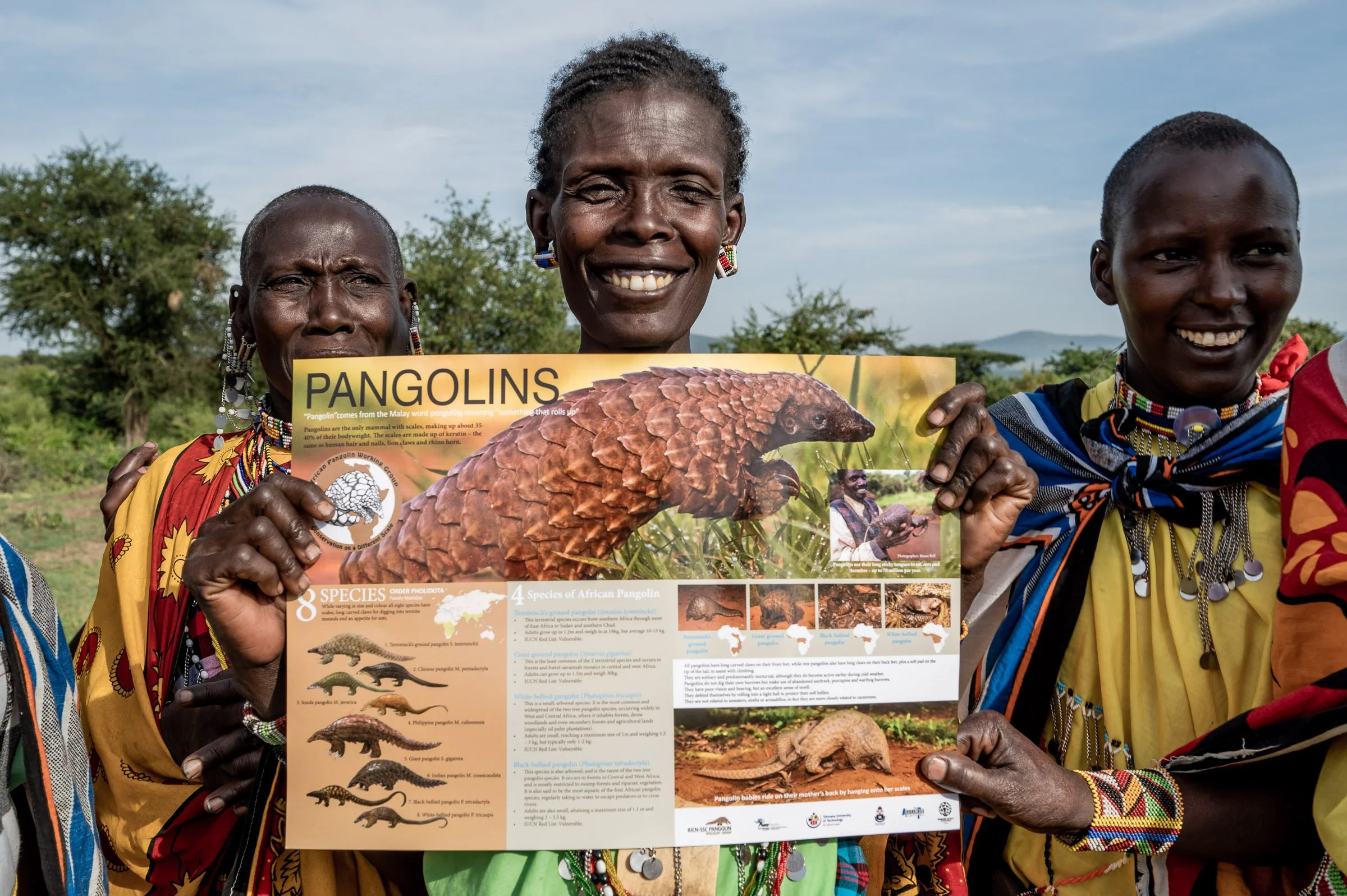 Three smiling women dressed in colorful traditional clothing holding a pamphlet about pangolins outdoors with trees and a cloudy sky in the background.