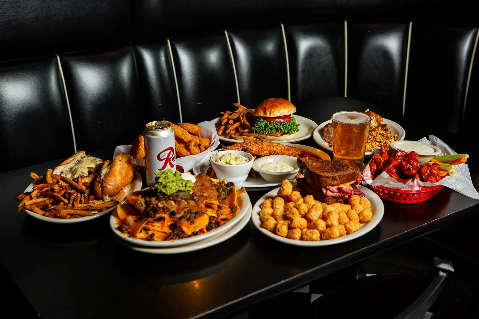 A spread of various American comfort foods on a black table, including sandwiches, fried chicken, pasta, tater tots, nachos, and a bowl of buffalo wings, with a can of beer and a draft pint.
