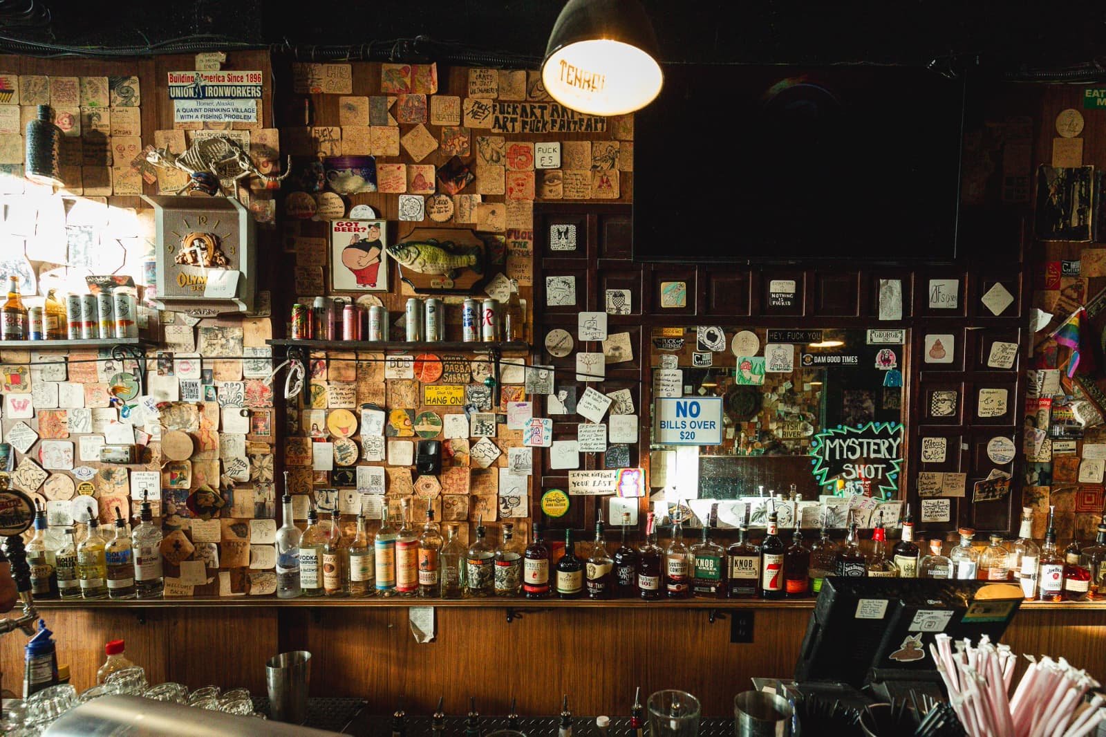 Bar interior with a brick wall decorated with various stickers, signs, and artwork, featuring a large mirror, bottles of liquor on the counter, and signs including 'NO BILLS OVER $20' and 'MYSTERY SHOT'