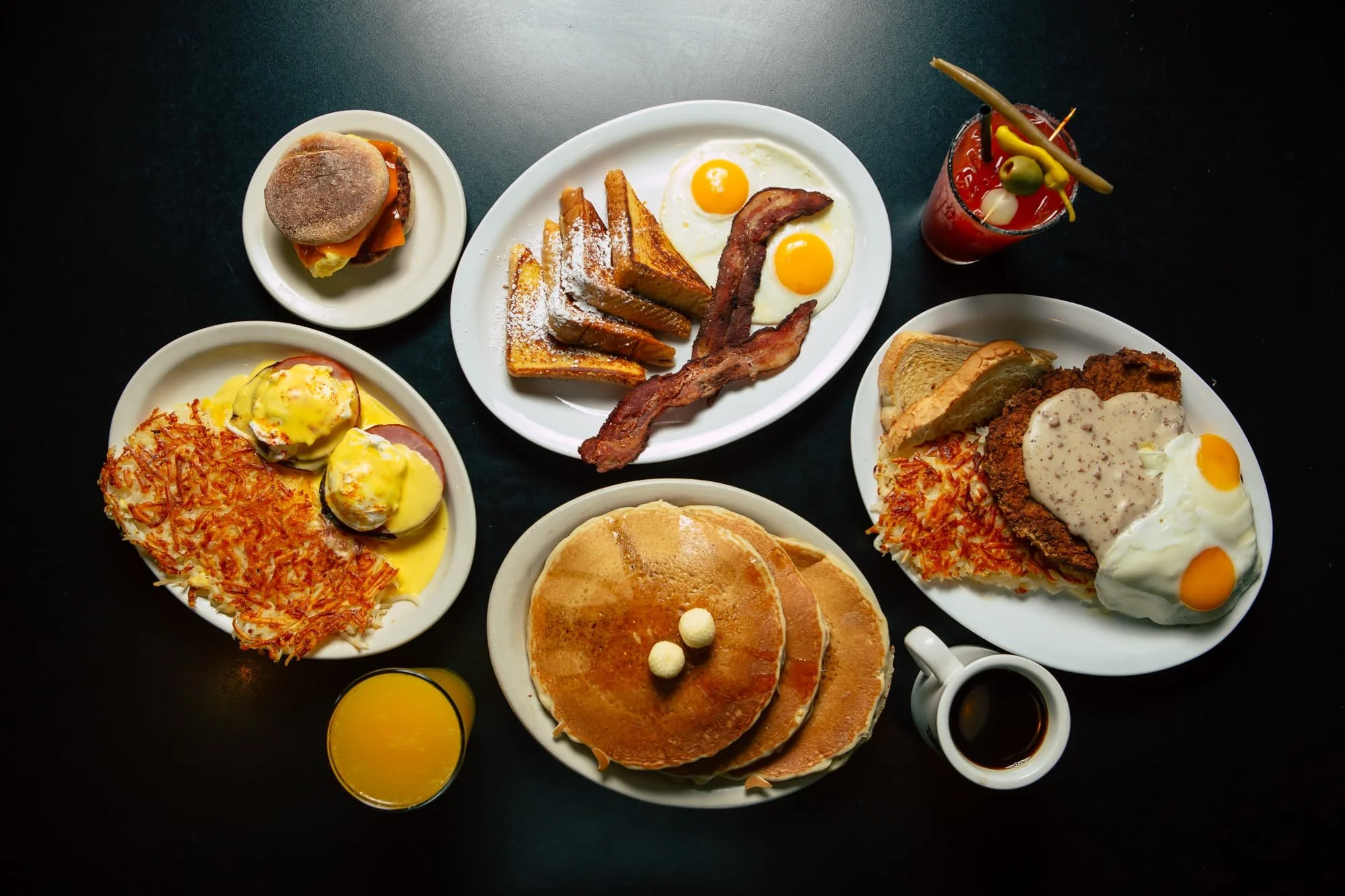 A breakfast spread with pancakes, eggs, bacon, toast, hash browns, a breakfast sandwich, muffins, fruit juice, and a Bloody Mary on a dark table.