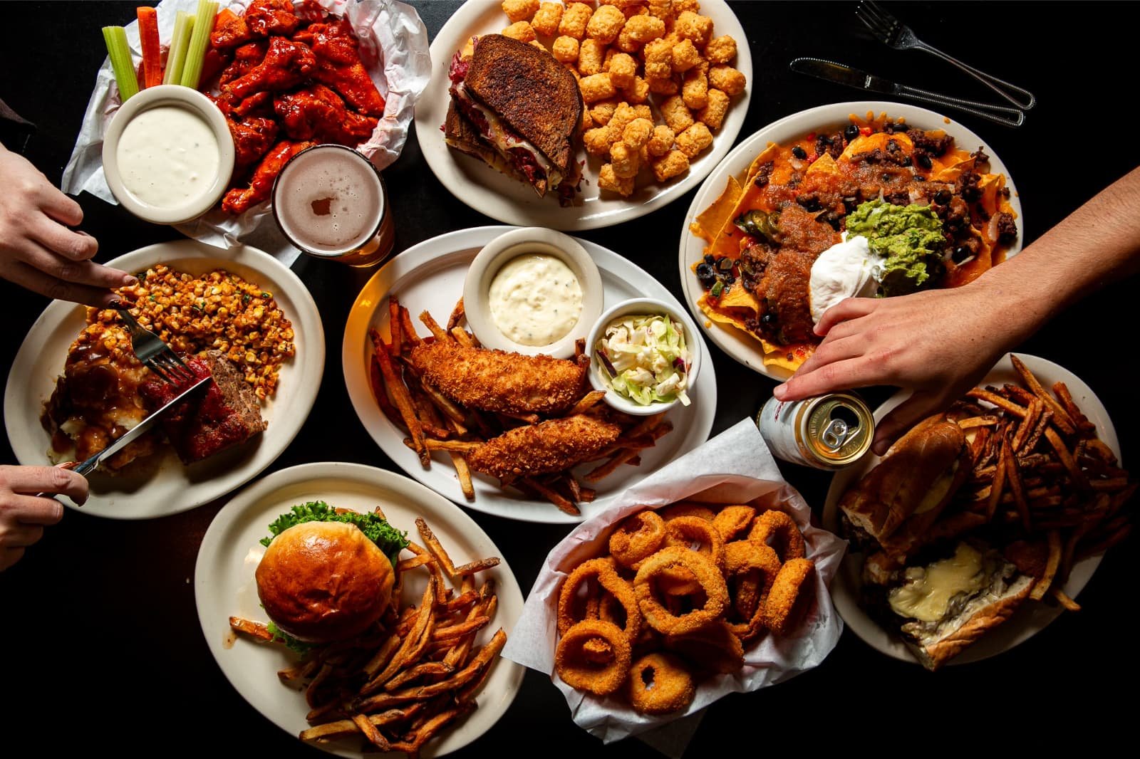 A table filled with various plates of American comfort food, including fried chicken, onion rings, cheeseburgers, nachos, pasta with sauce, ribs, and chicken wings, alongside drinks and condiments.