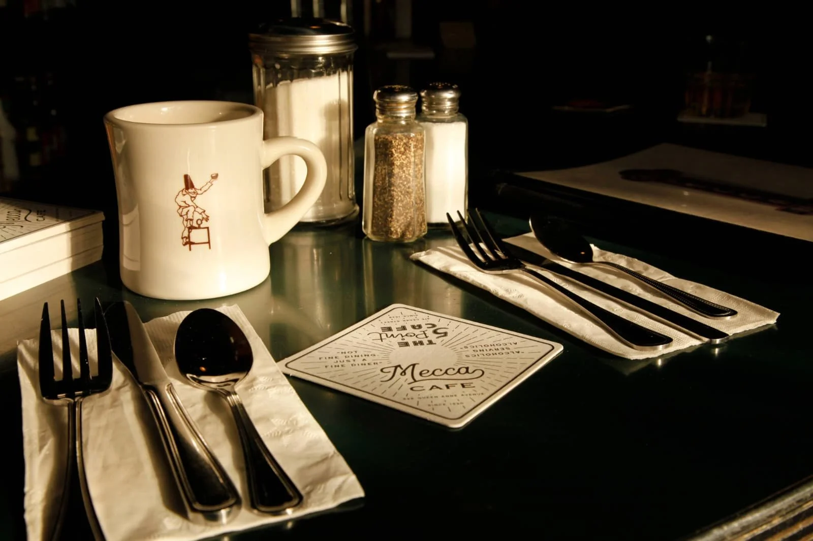 Table setting at a cafe with a white mug featuring a red illustration, a napkin with cutlery, and condiments in glass dispensers, with a menu card that reads 'Mecca Cafe'.