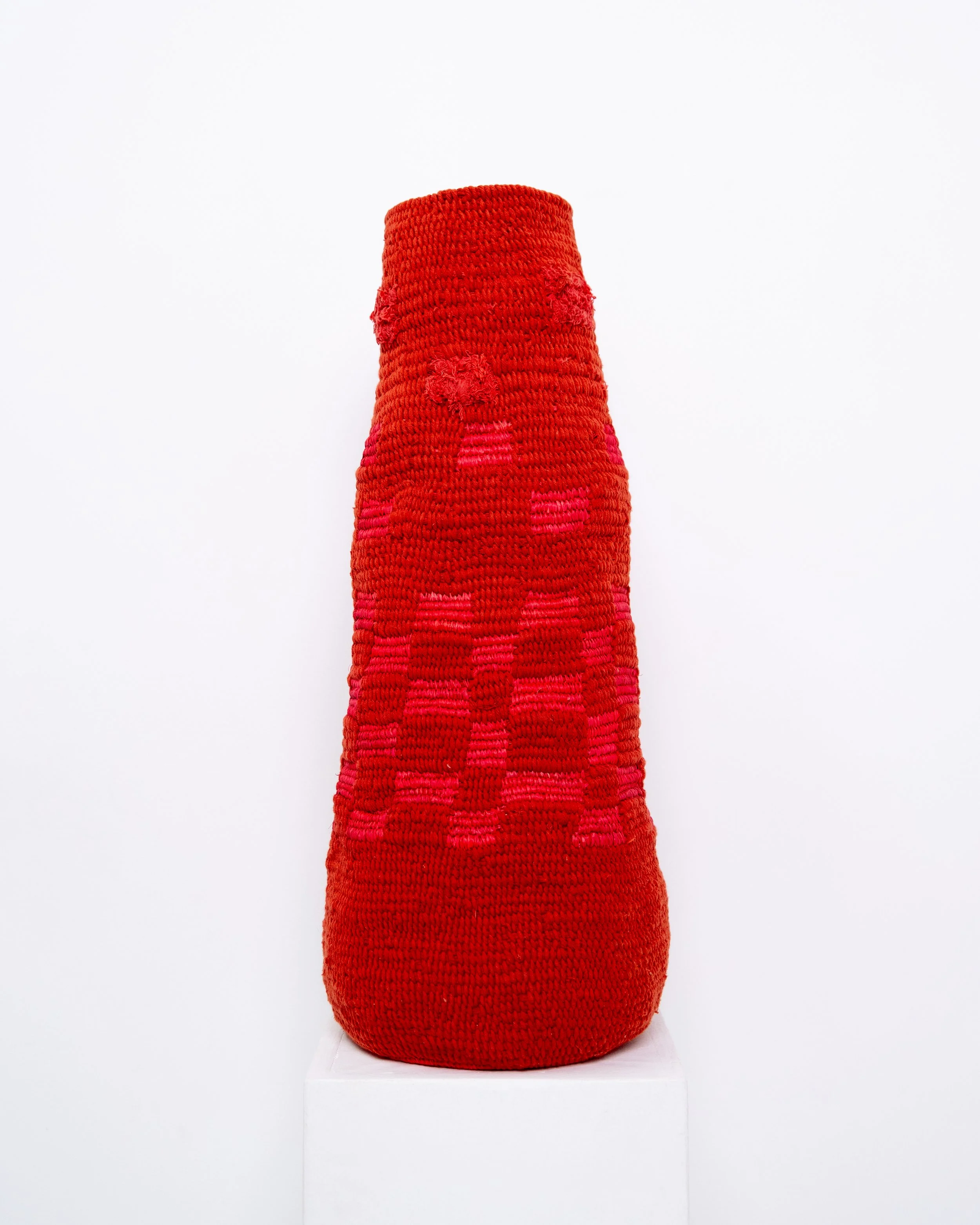 Red and pink woven textile art piece displayed on a white pedestal against a plain white background.