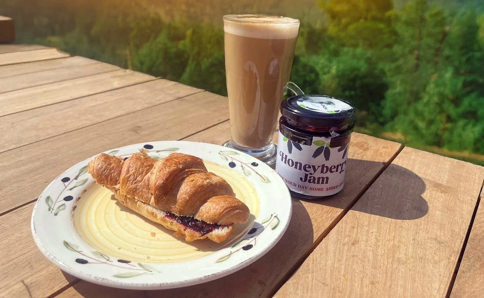 A plate with a croissant and honeyberry jam, a tall glass of coffee or hot chocolate, and a jar of honeyberry jam on a wooden table outdoors