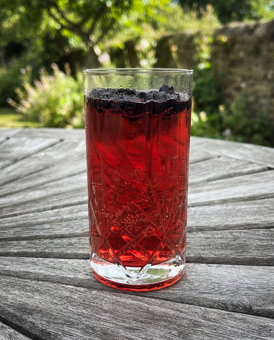 A tall glass filled with a red drink and ice cubes, placed on a wooden table outdoors.
