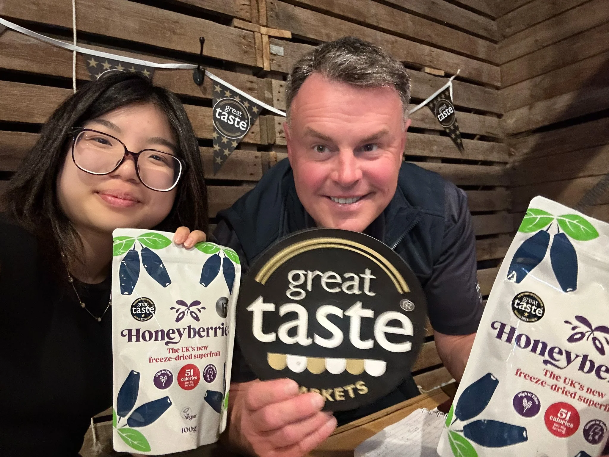 Two people smiling, holding away packages of frozen honeyberries with a wooden wall background and bunting flags. One person is a young woman with glasses, and the other is a man with short hair. They are celebrating with a branded sign that reads 'great taste'.