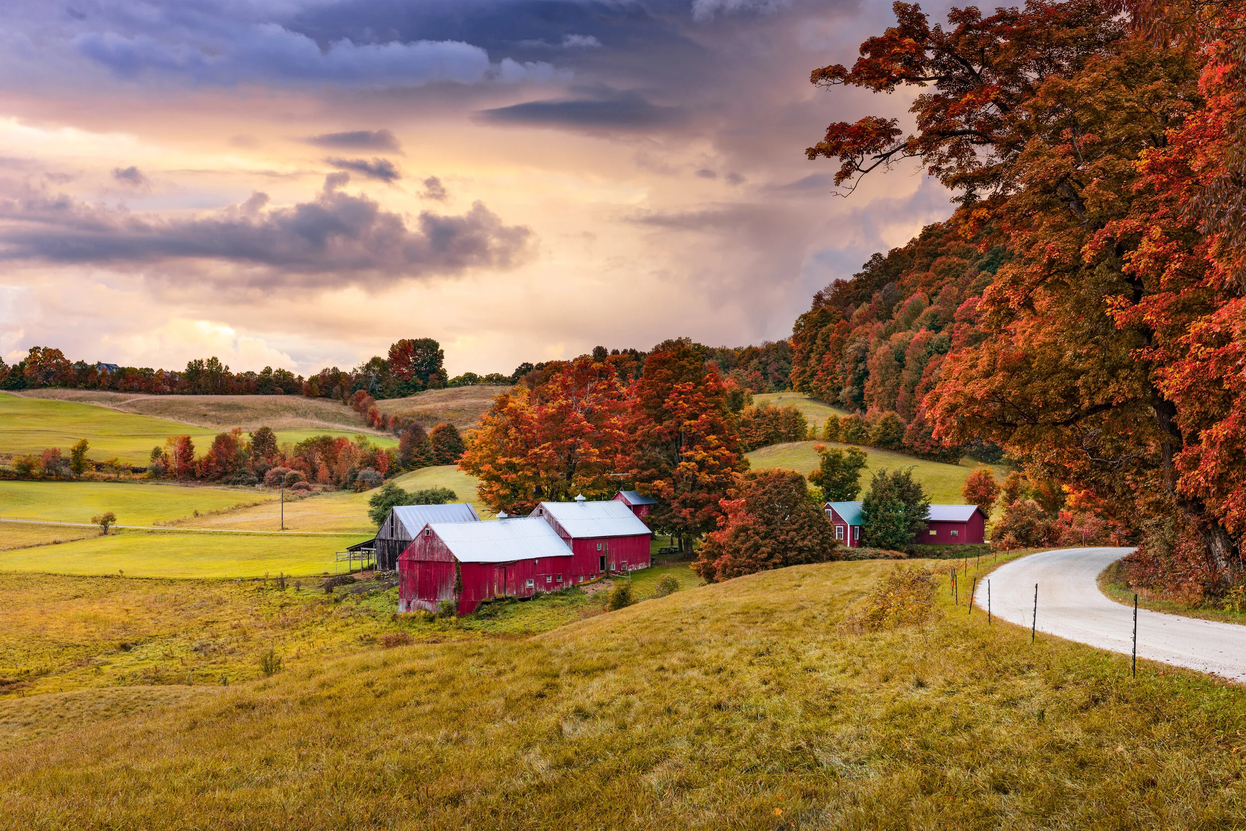 A scenic countryside landscape featuring rolling hills, red barns, and colorful autumn trees under a partly cloudy sky.