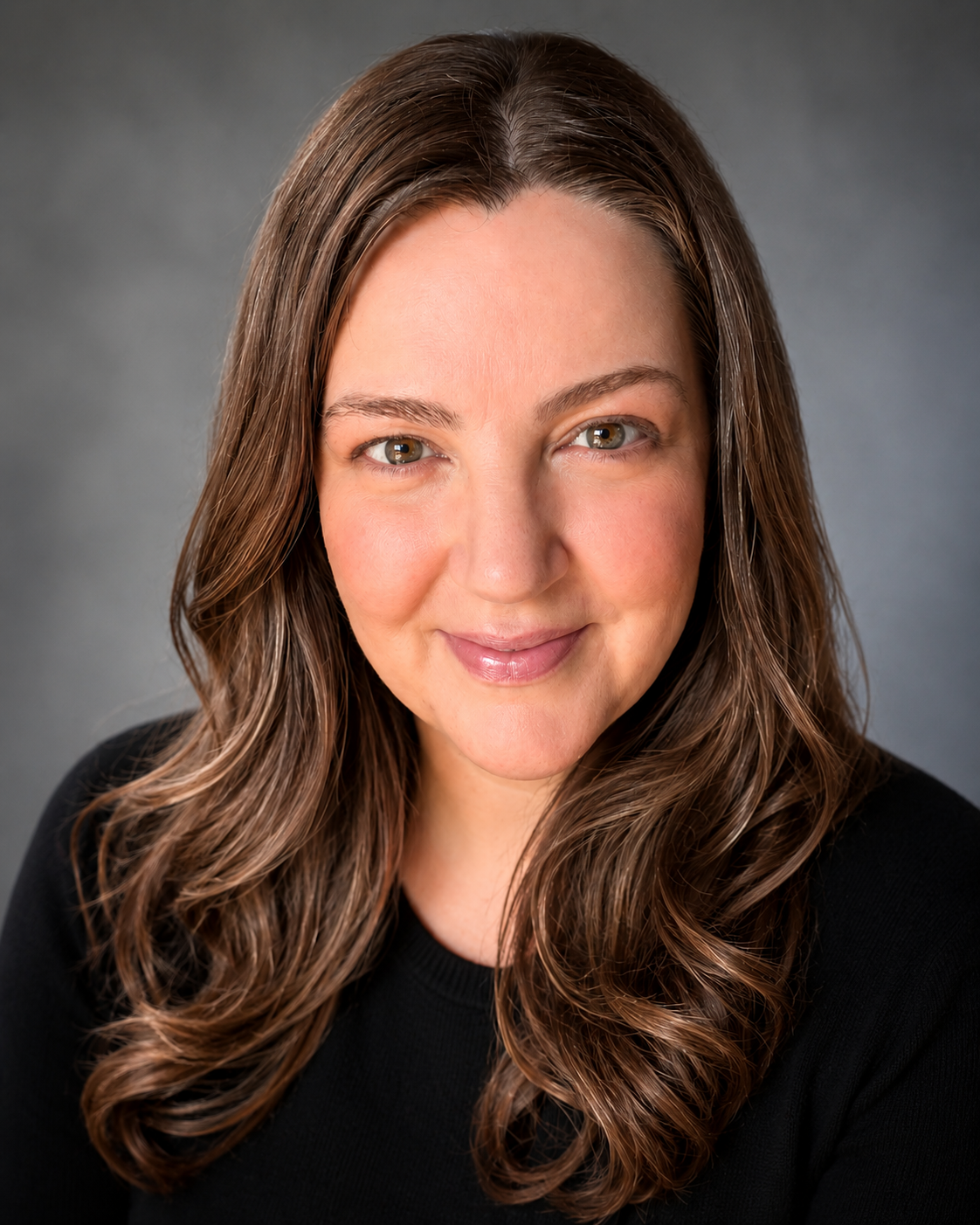 A woman with long wavy brown hair, light skin, and green eyes smiling in a black top against a gray background.