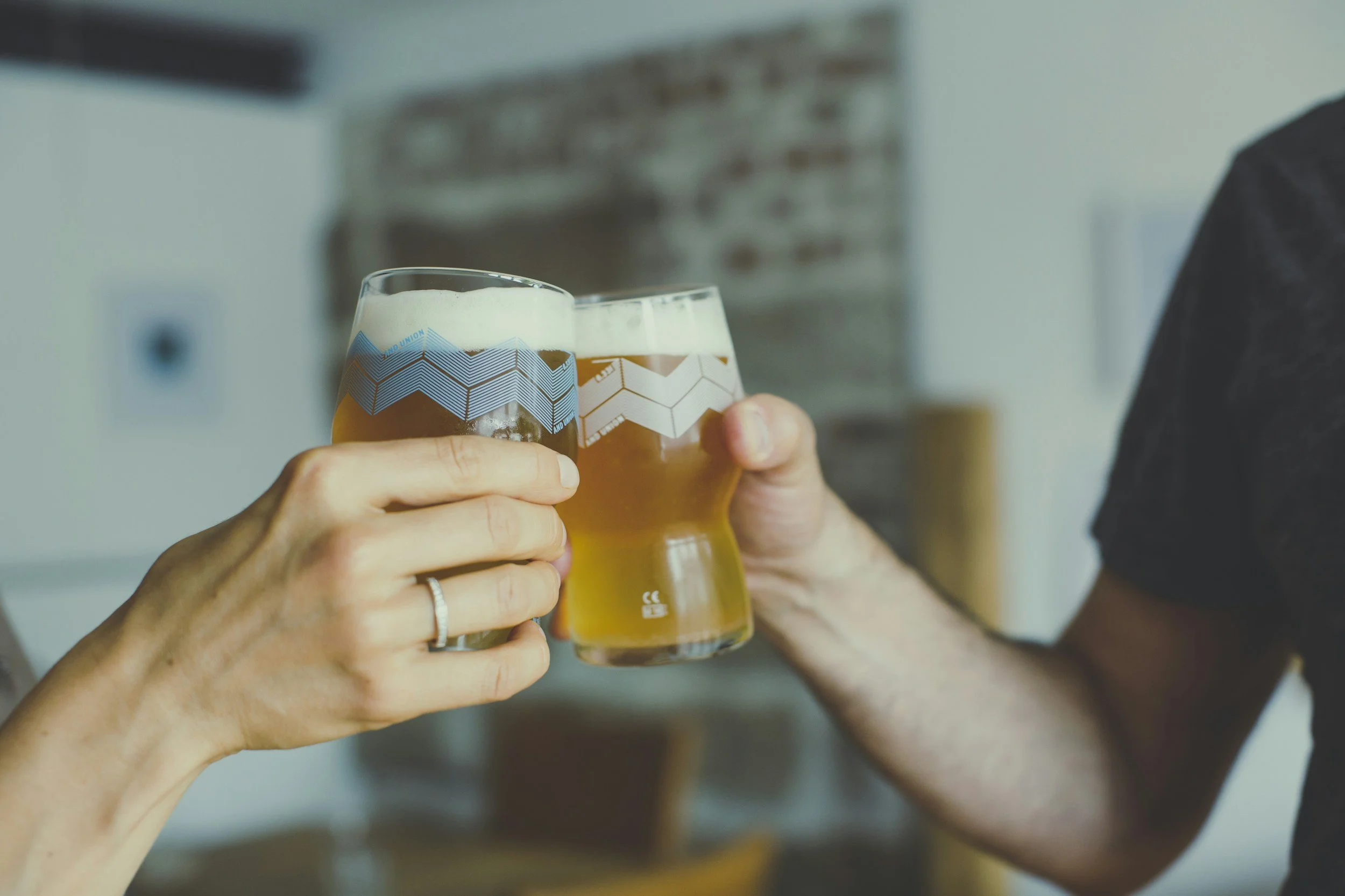 Two people clinking glasses of beer in a toast, close-up of hands with a blurred indoor background.