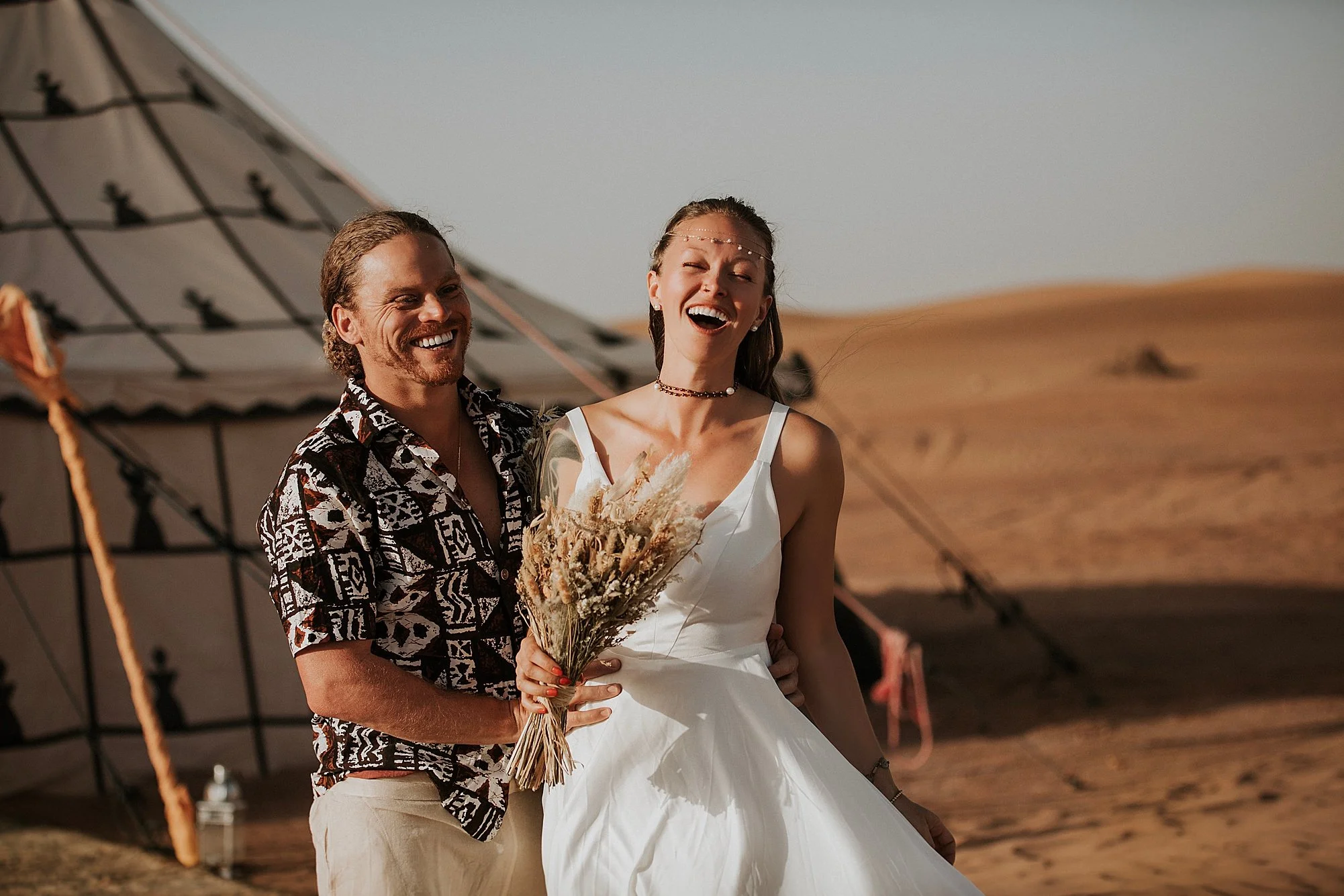 a woman and her husband laughing in their wedding day and standing in front of their luxurious tent in moroccan sahara desert