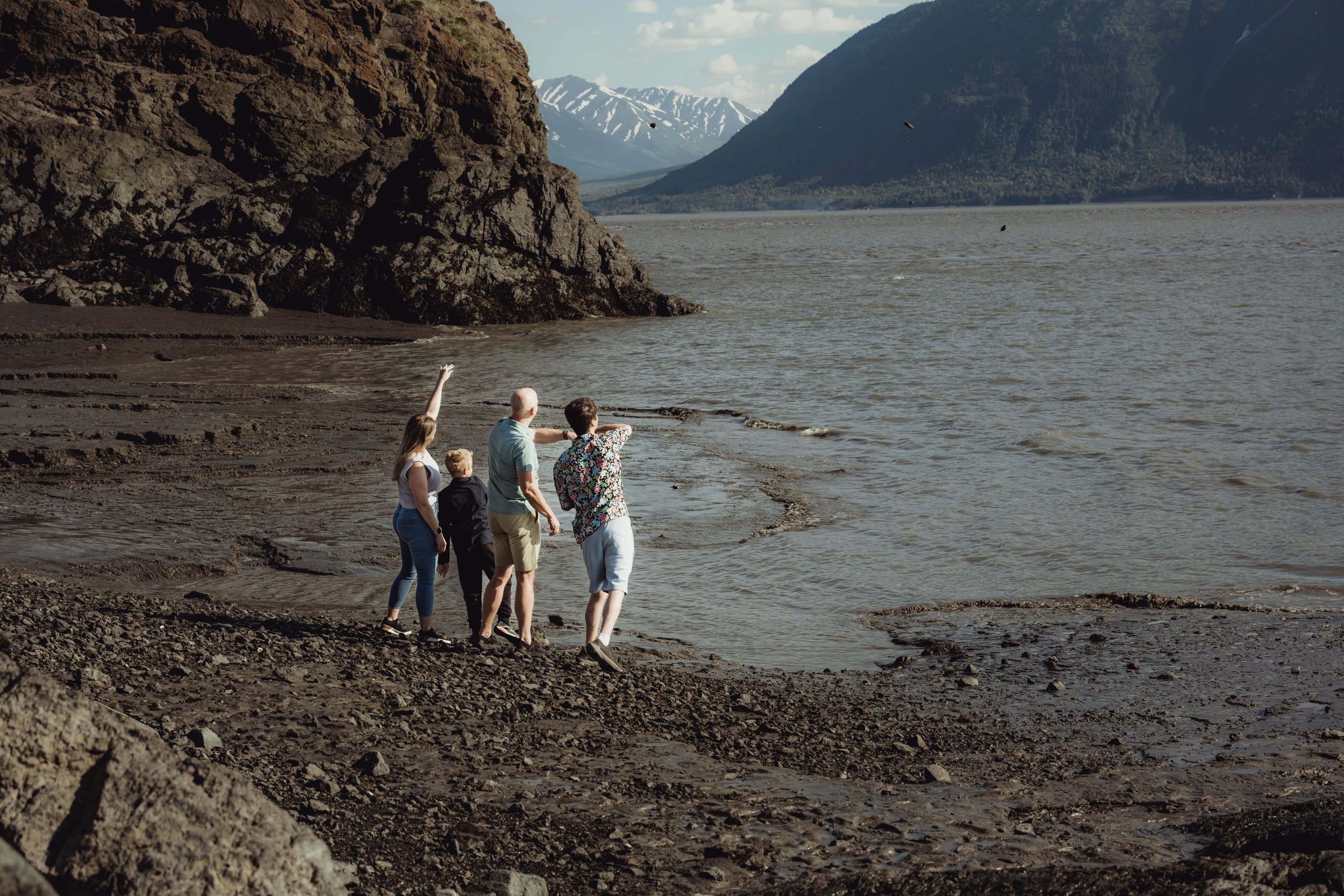 Five people walking along a rocky shoreline by a large body of water, with mountainous terrain in the background.
