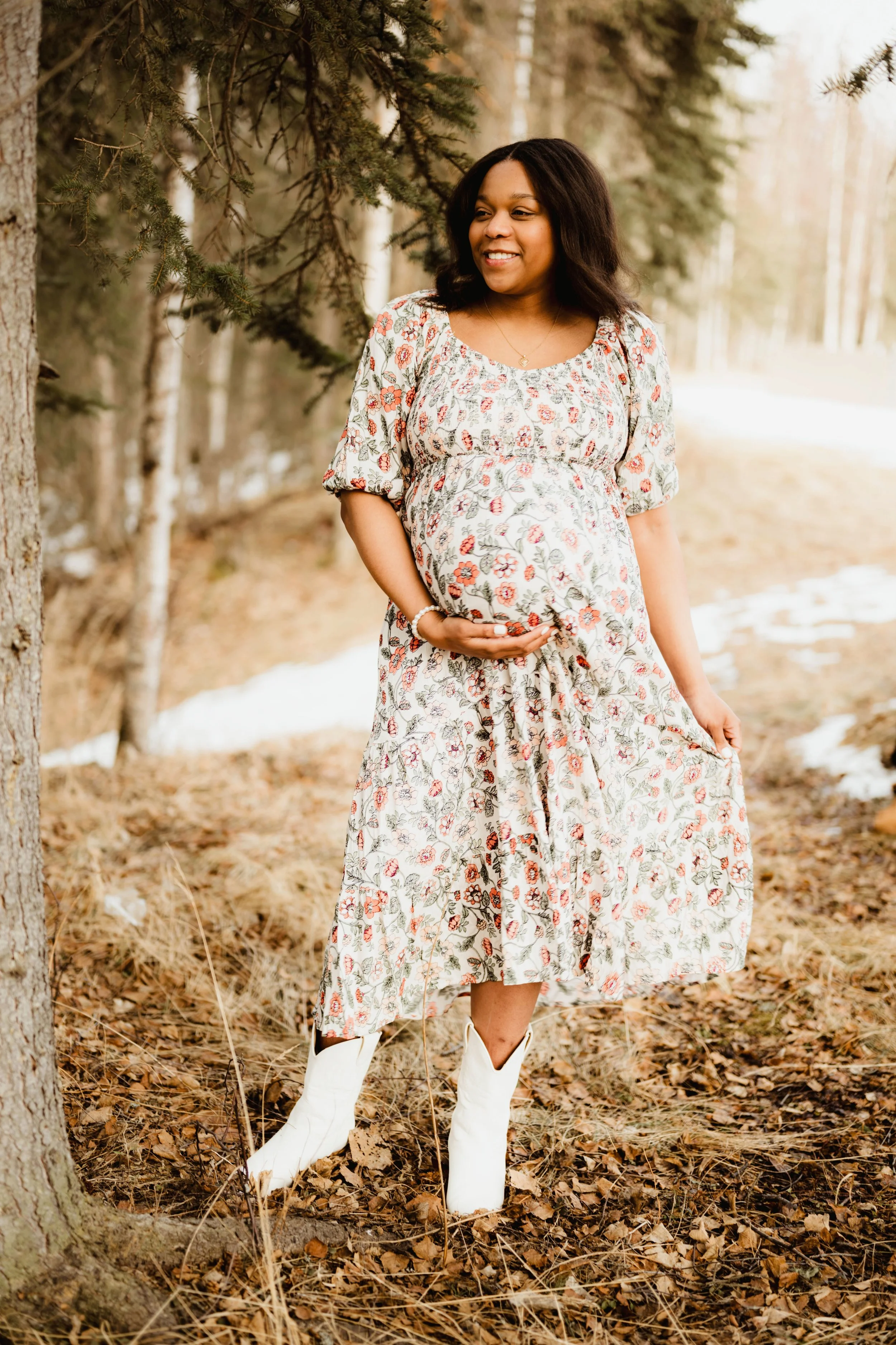 A pregnant woman in a floral dress and white cowboy boots standing outdoors among trees and fallen leaves, touching her belly and smiling.