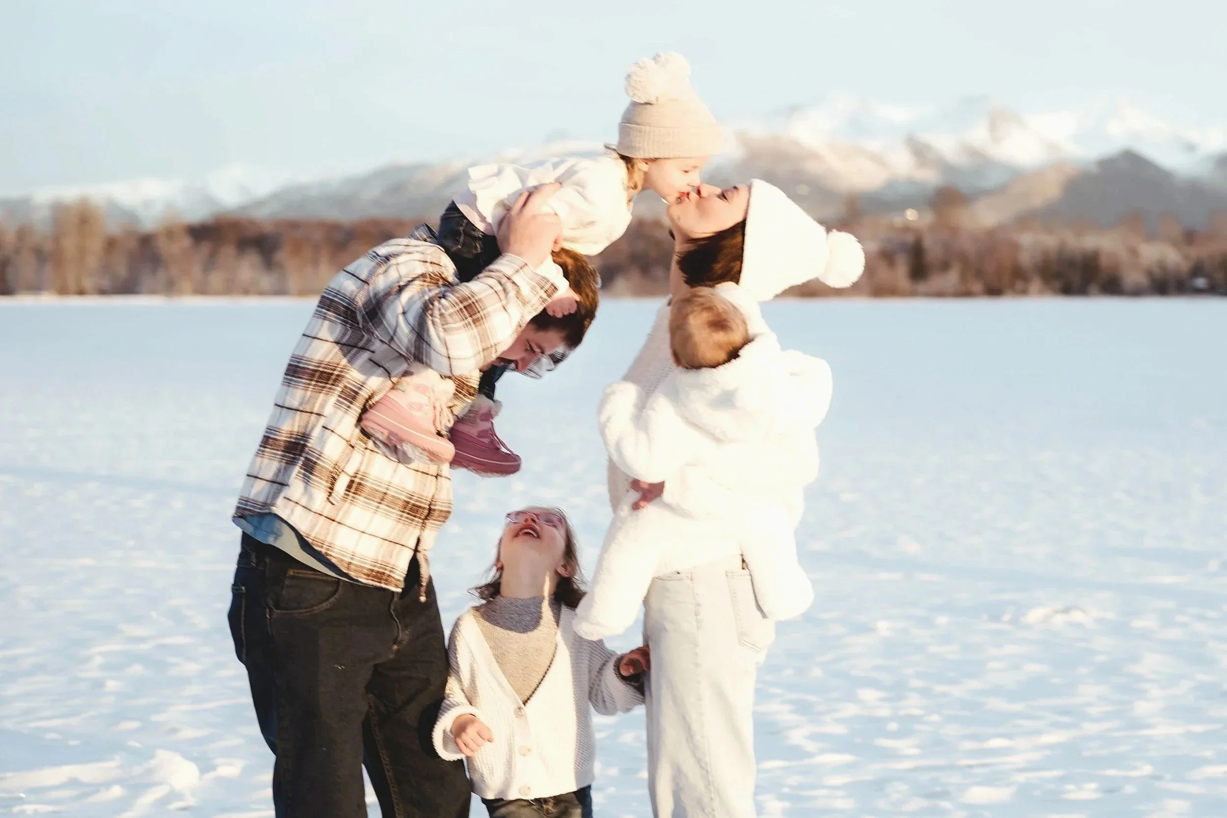 A family of five outside in the snow, with mountains in the background. They are wearing winter clothes, and the two adults are lifting two children in the air while the others look up and laugh.