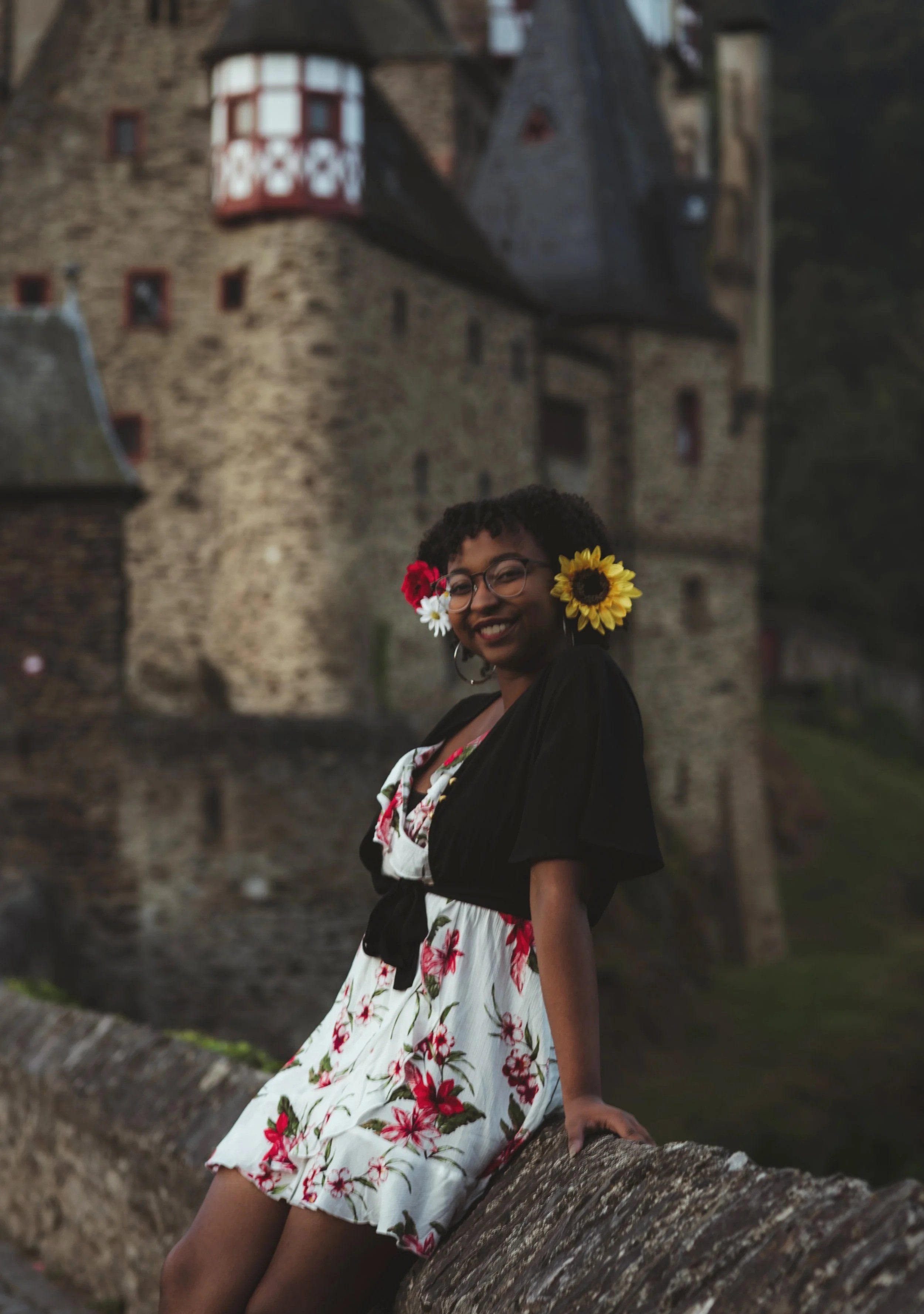 A young woman with glasses and flowers in her hair, wearing a floral dress, sitting on a stone ledge in front of a castle, smiling.