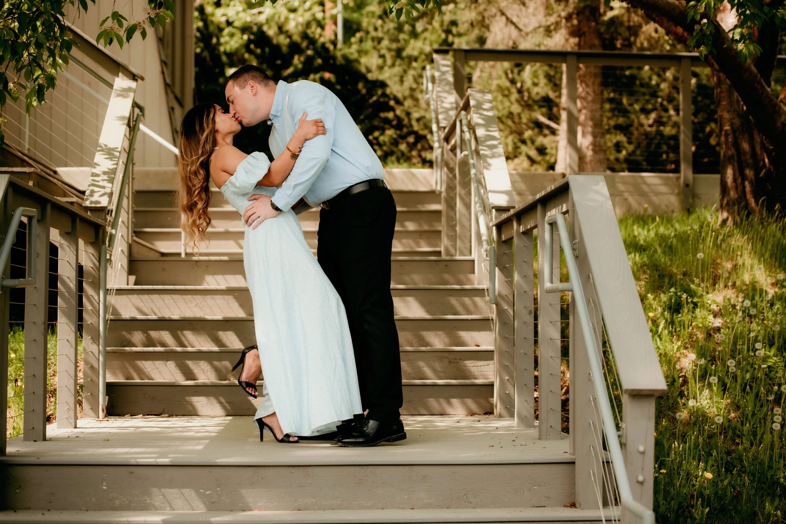 A couple sharing a kiss on outdoor stairs, with the woman wearing a light blue dress and high heels, and the man in a light blue shirt and black pants, surrounded by green trees and grass.