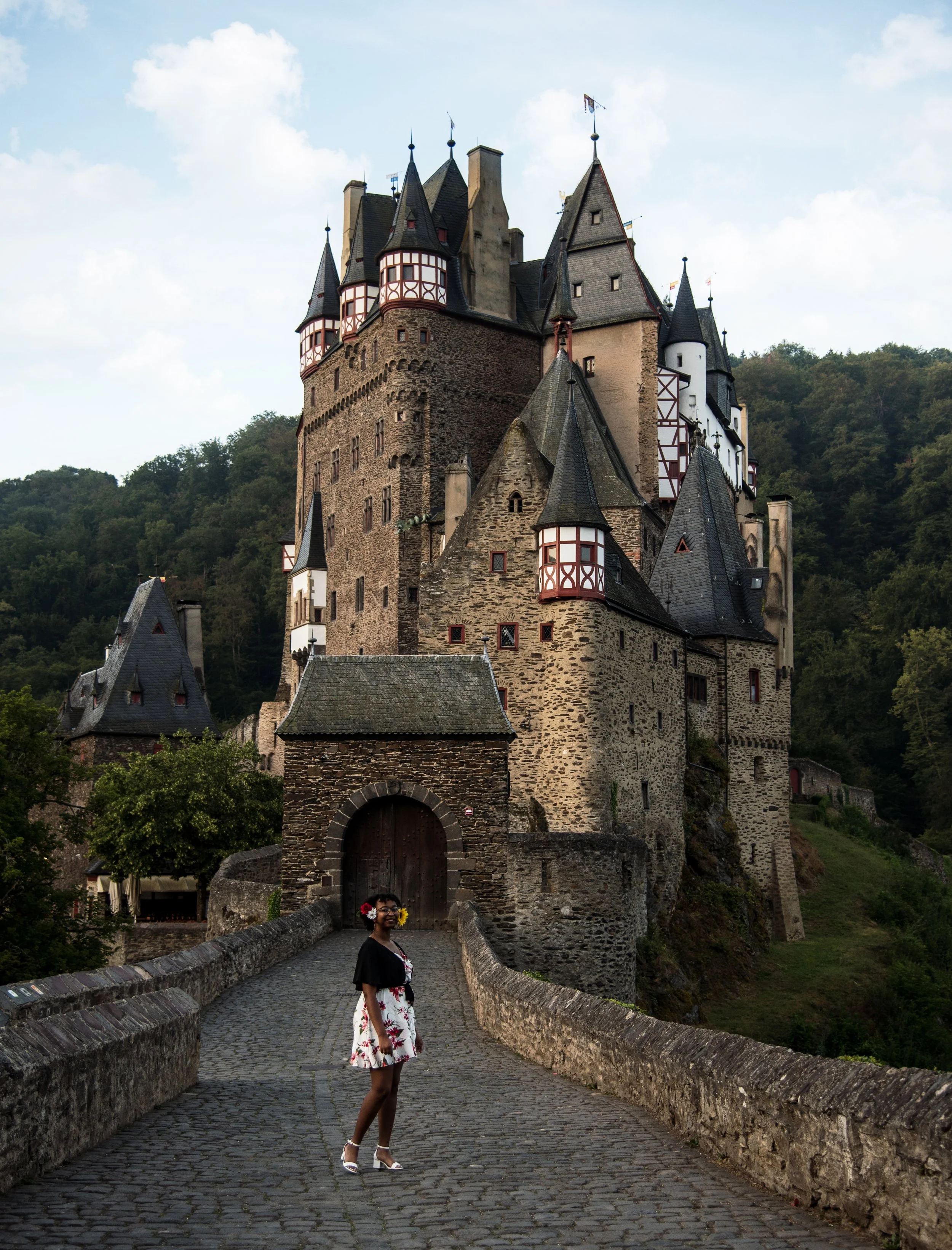 A young woman stands on a cobblestone bridge in front of a medieval stone castle with multiple towers, set against a wooded hillside under partly cloudy sky.
