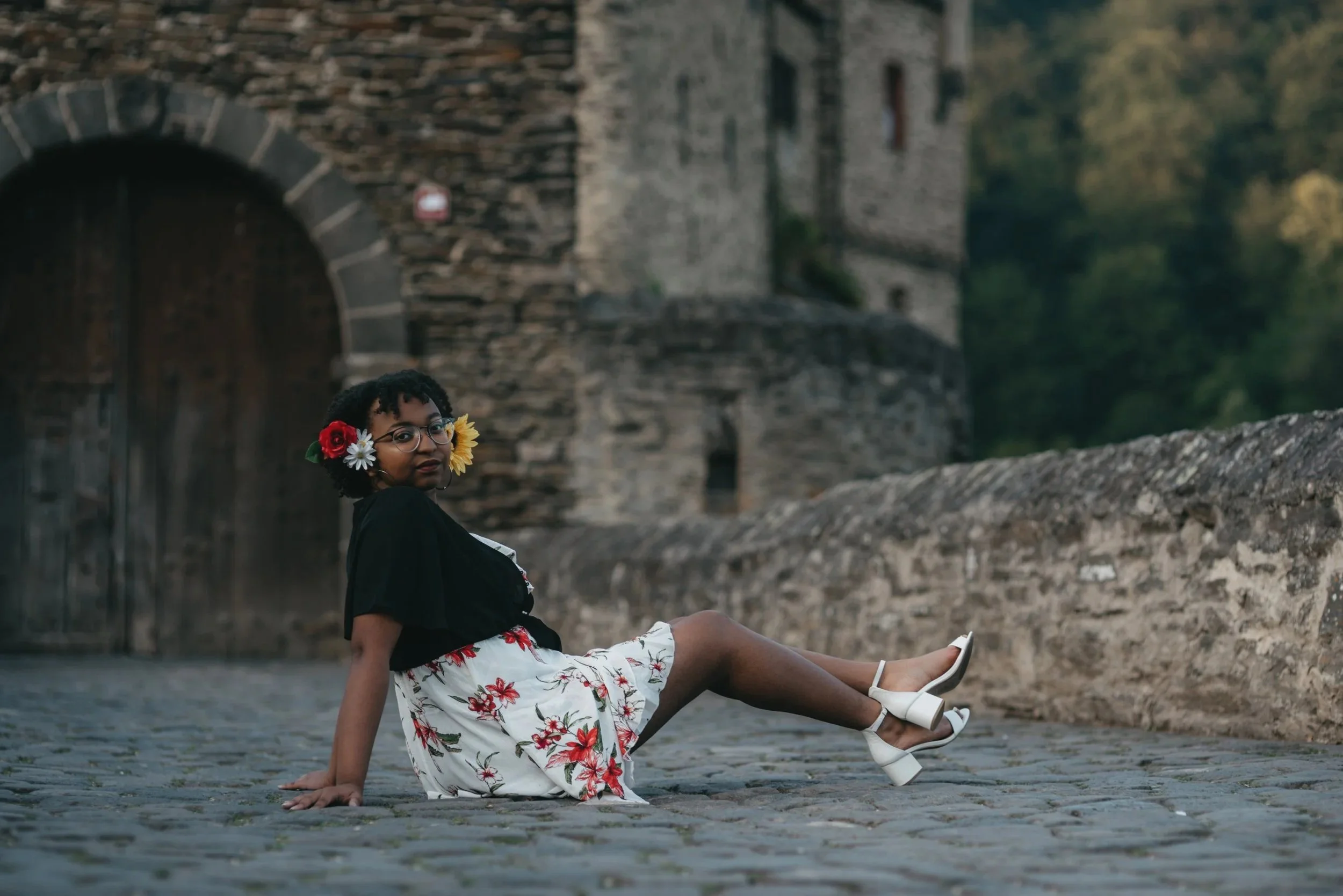 A woman with short curly hair, glasses, and flowers in her hair, sitting on a cobblestone street near an old stone building with an arched wooden door, dressed in a floral skirt, black top, and white heels.