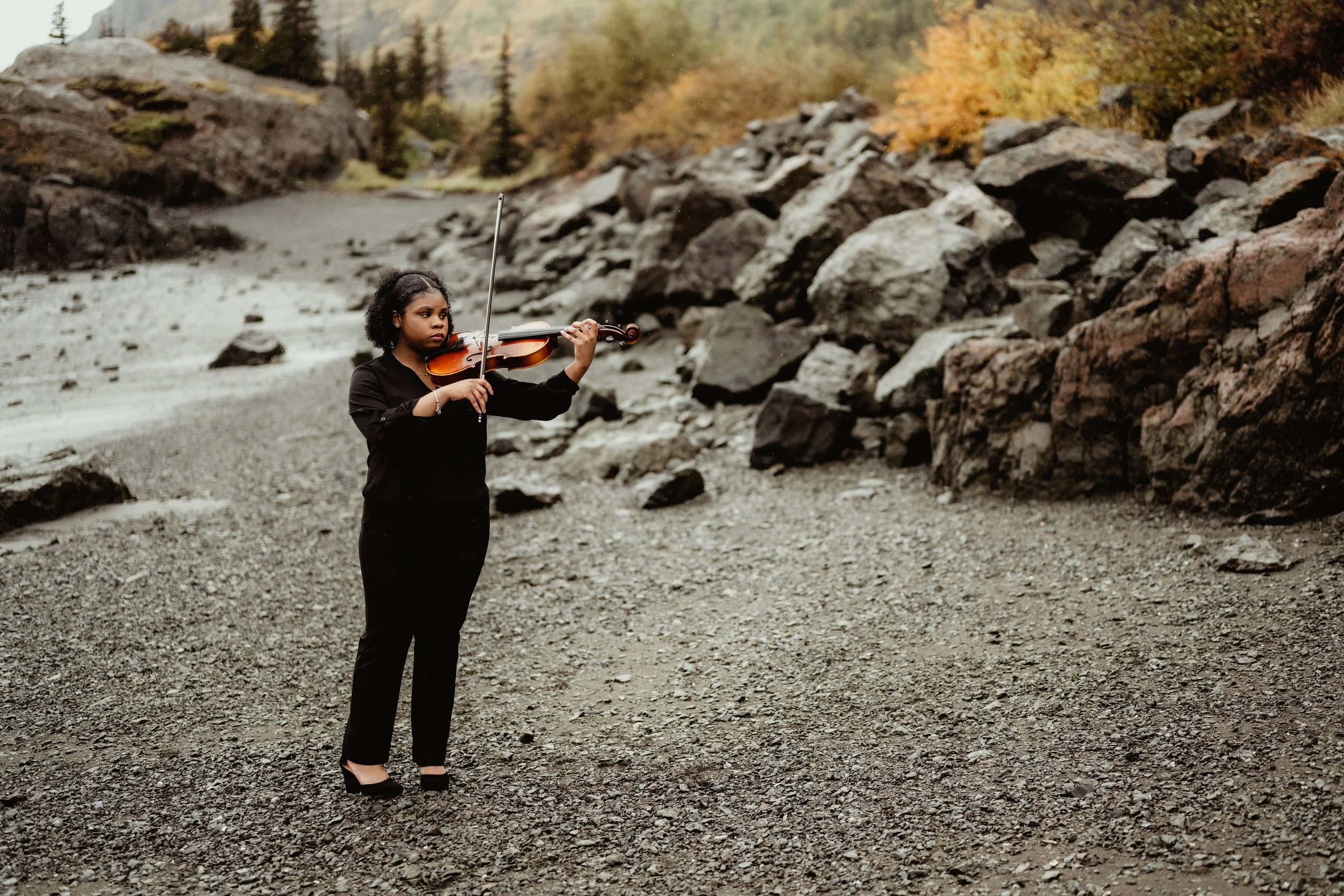 A woman playing the violin outdoors on rocky terrain with a river and autumn trees in the background.