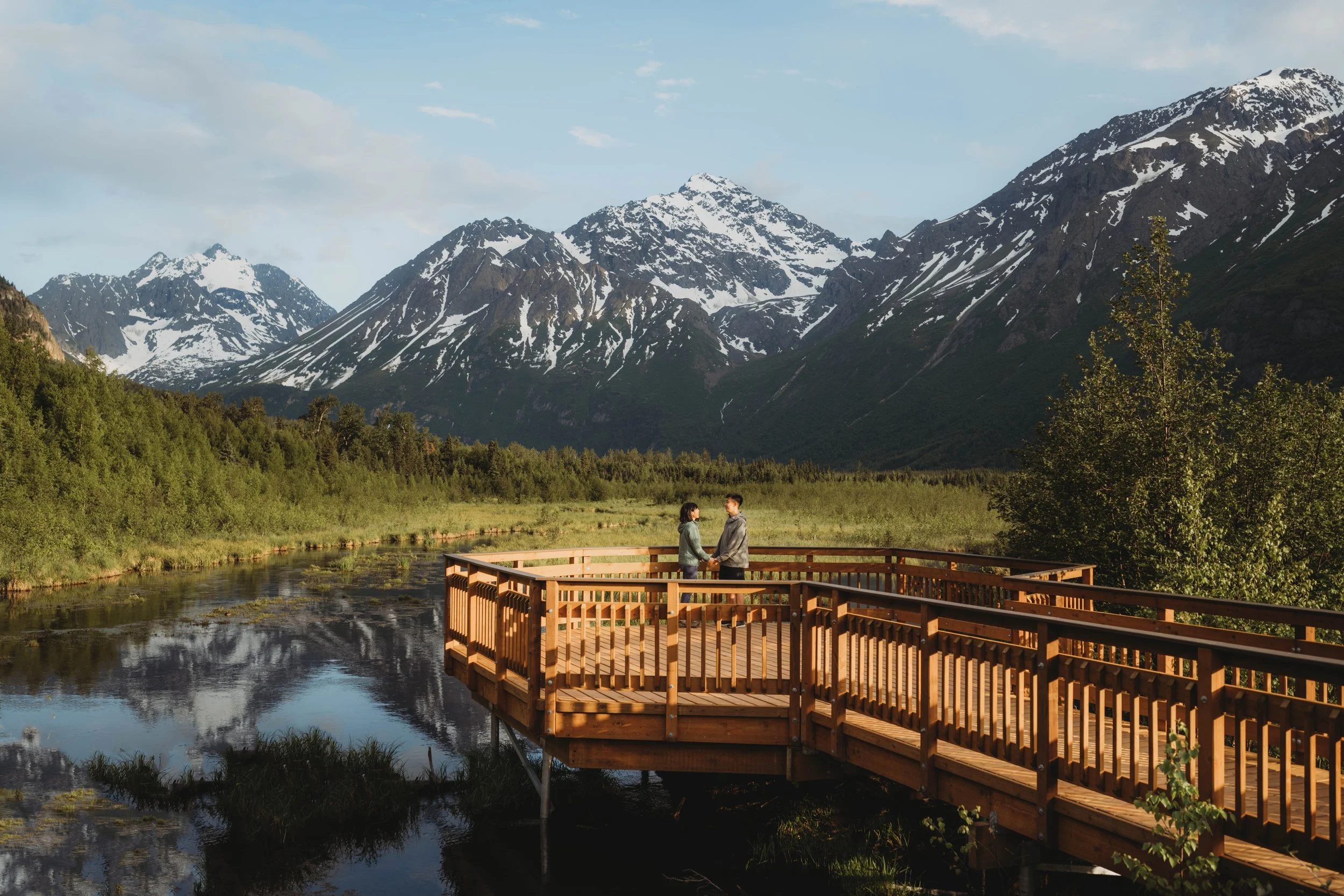 Two people holding hands on a wooden viewing platform overlooking a river with snow-capped mountains in the background.