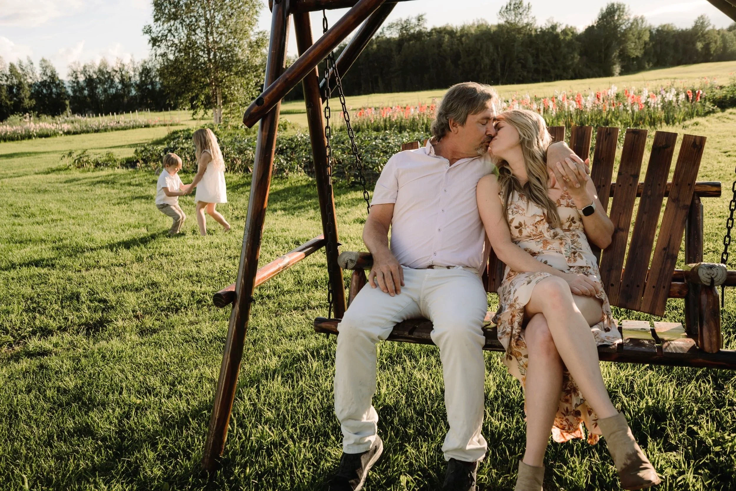 A couple sharing a kiss on a wooden swing outdoors on a sunny day, with two children playing nearby in a grassy field with flowers and trees in the background.
