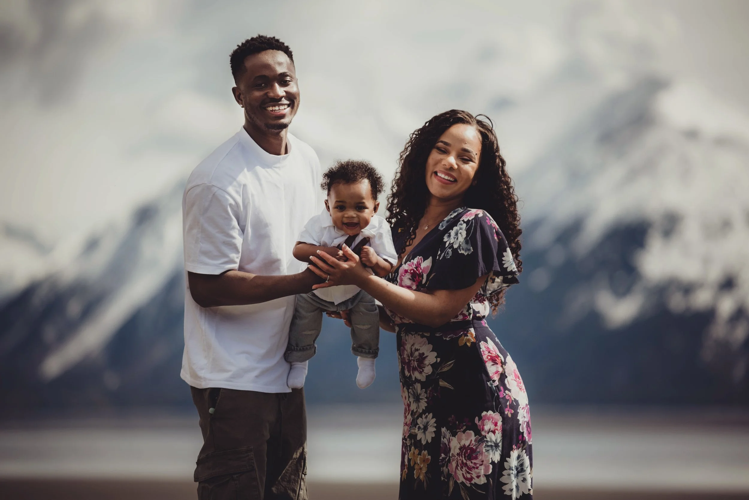 A happy family of three standing outdoors with snow-capped mountains in the background, holding and smiling at their young toddler.