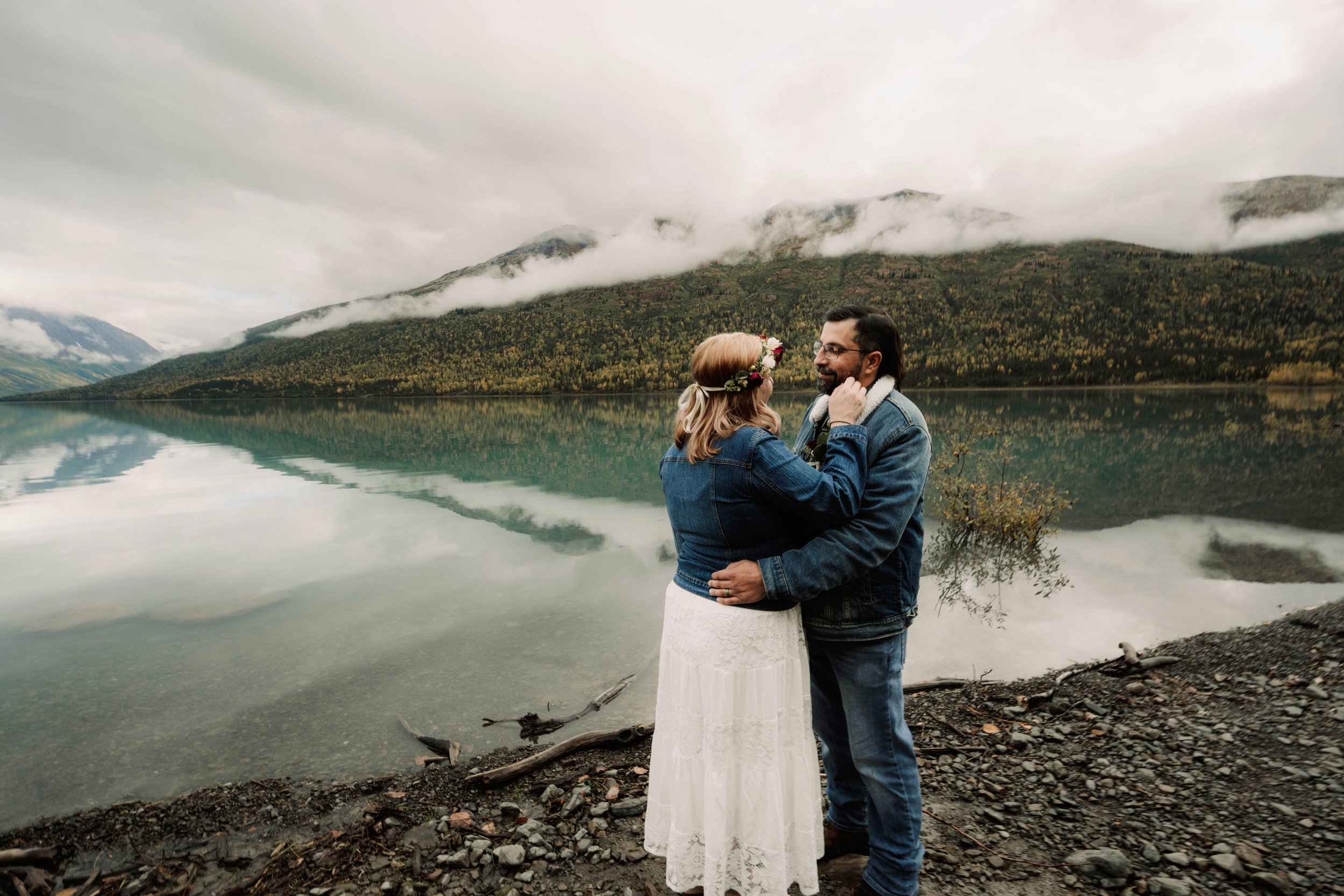 A couple embracing by a lake with mountains in the background during cloudy weather, woman wearing a flower crown and white dress, man in denim jacket and jeans.