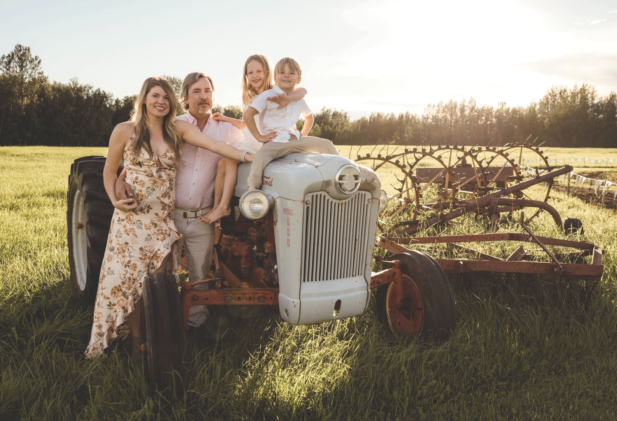 A family of four standing on and around an old tractor in a grassy field during sunset. Two children sit on the tractor, while a woman and man stand beside it.