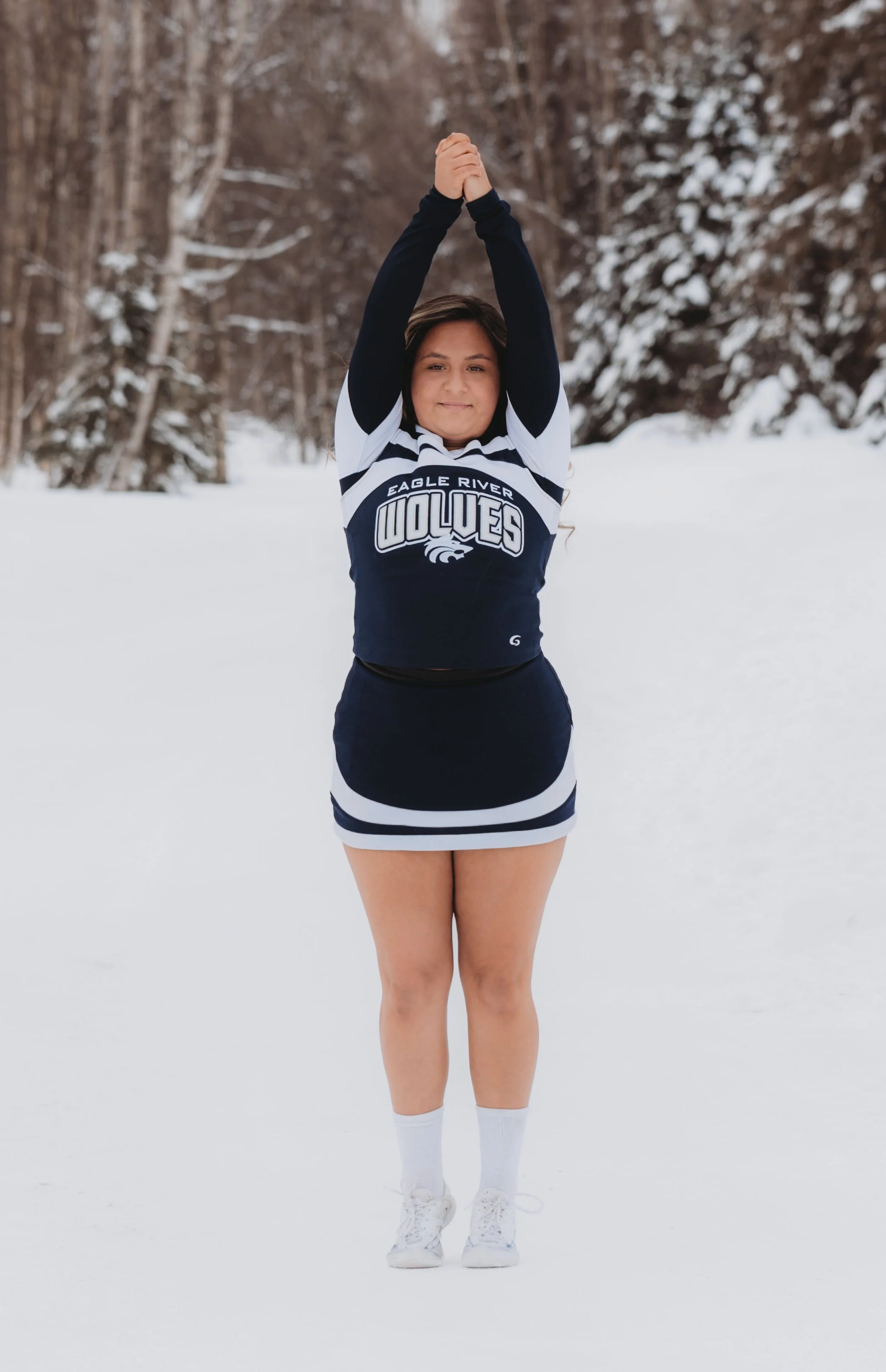 A young woman in a cheerleading uniform standing in the snow with arms stretched overhead, holding her hands together, with snow-covered trees in the background.